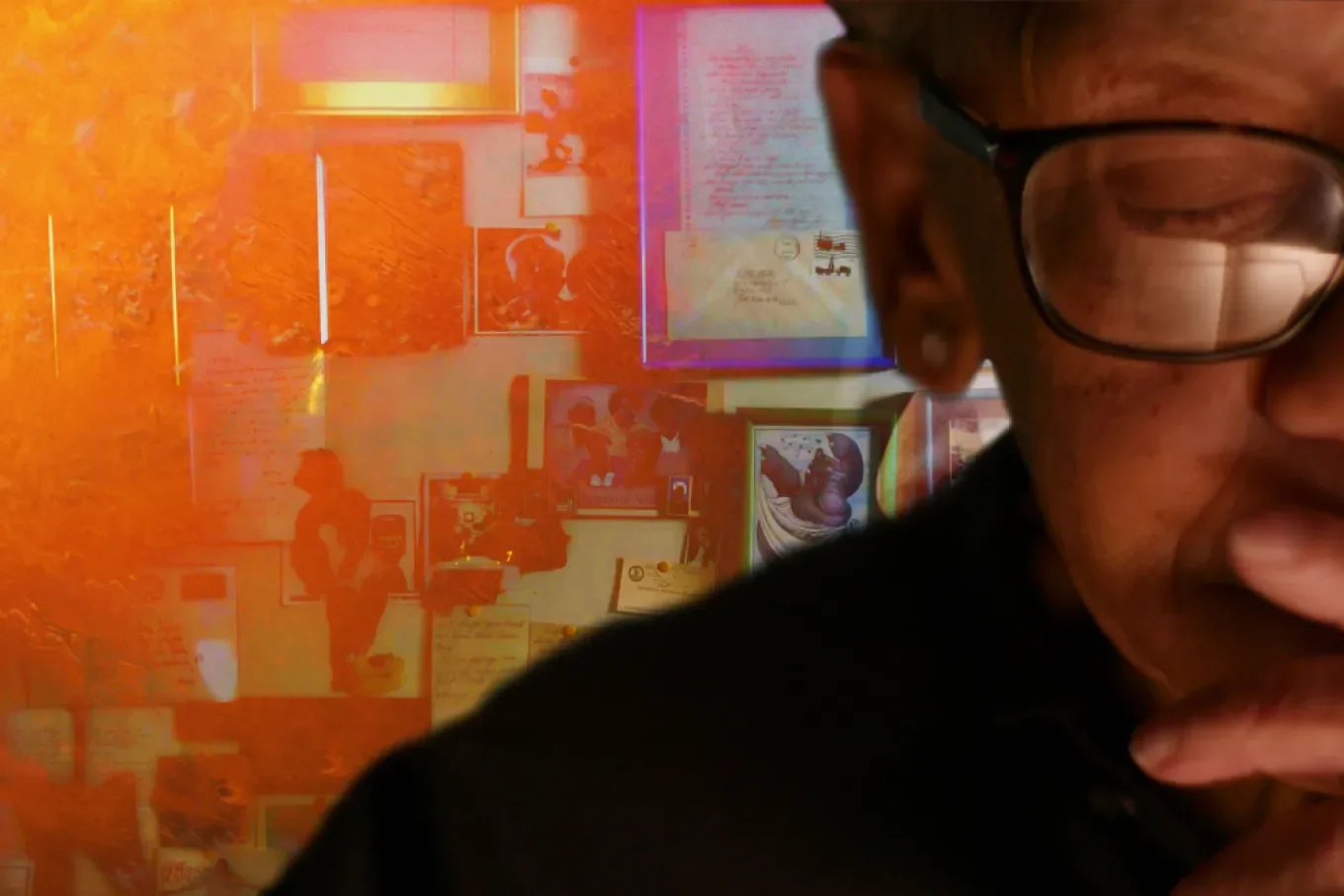 Close-up of a person with glasses and freckles, covering part of their face with their fingers, with a colorful collage of photos, drawings, and documents on a wall in the background.