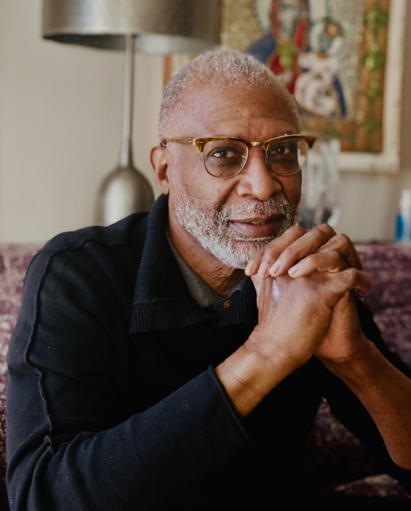 An elderly African American man with gray hair and beard, wearing glasses and a black sweater, sitting with hands clasped in front of his face, in a cozy indoor setting with artwork in the background.