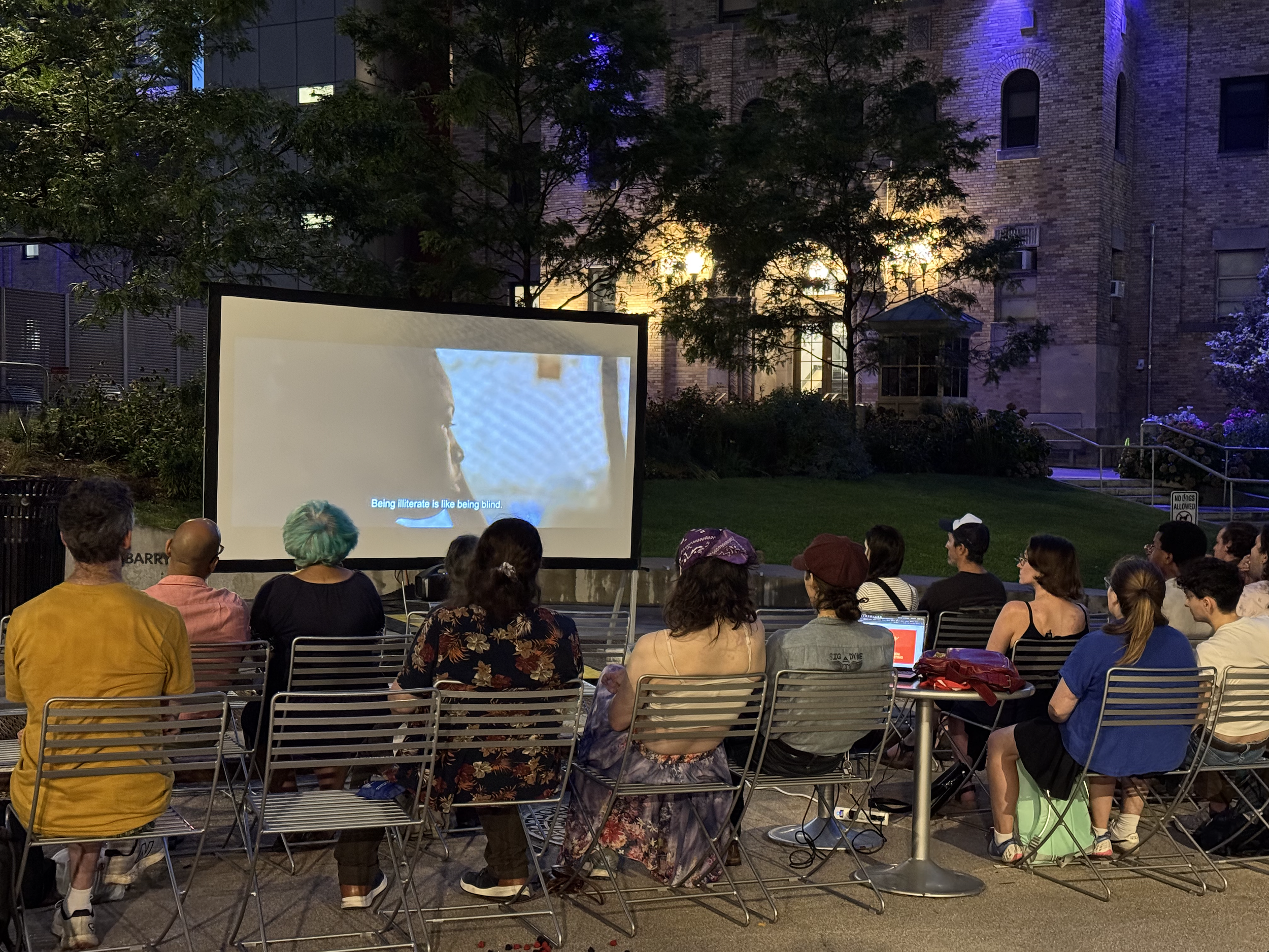 People watching a movie outdoors on a large screen in an urban park at night, with a building and trees illuminated in the background.