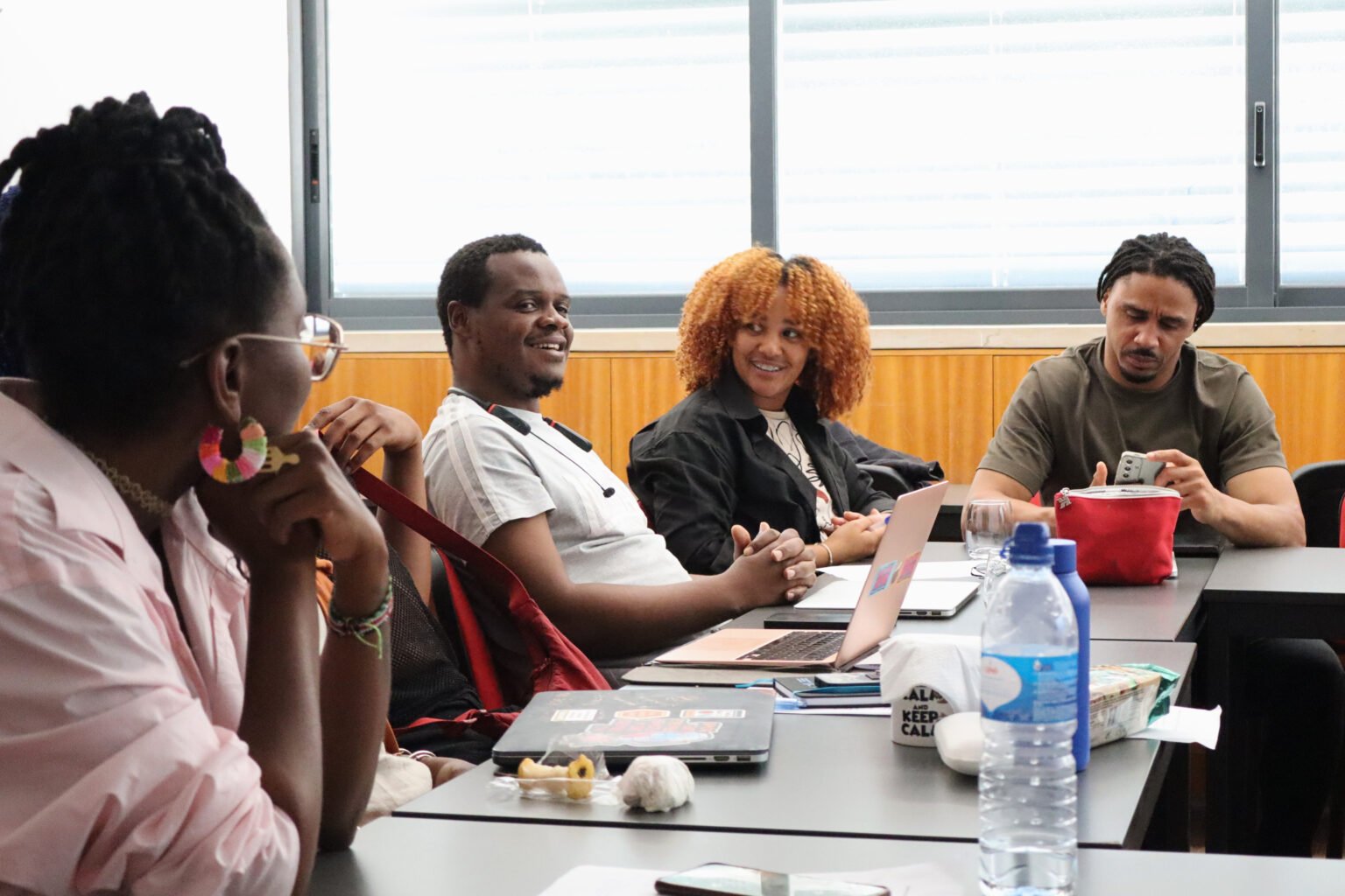 Four diverse individuals seated at a conference table in a meeting room, engaged in conversation with laptops and personal items on the table, and a window with blinds in the background.