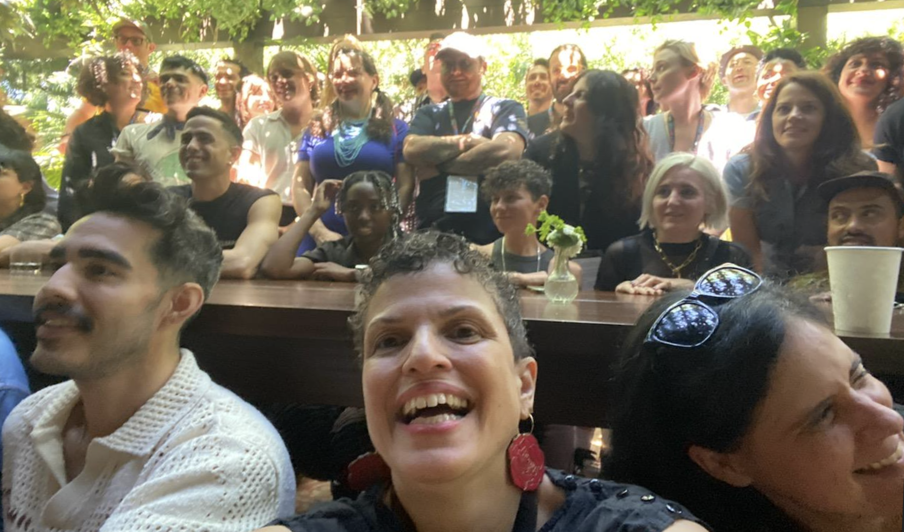 A group of smiling and happy people gathered outdoors, with some seated at a wooden table and others standing behind. The setting appears to be a sunny day with greenery and trees in the background.