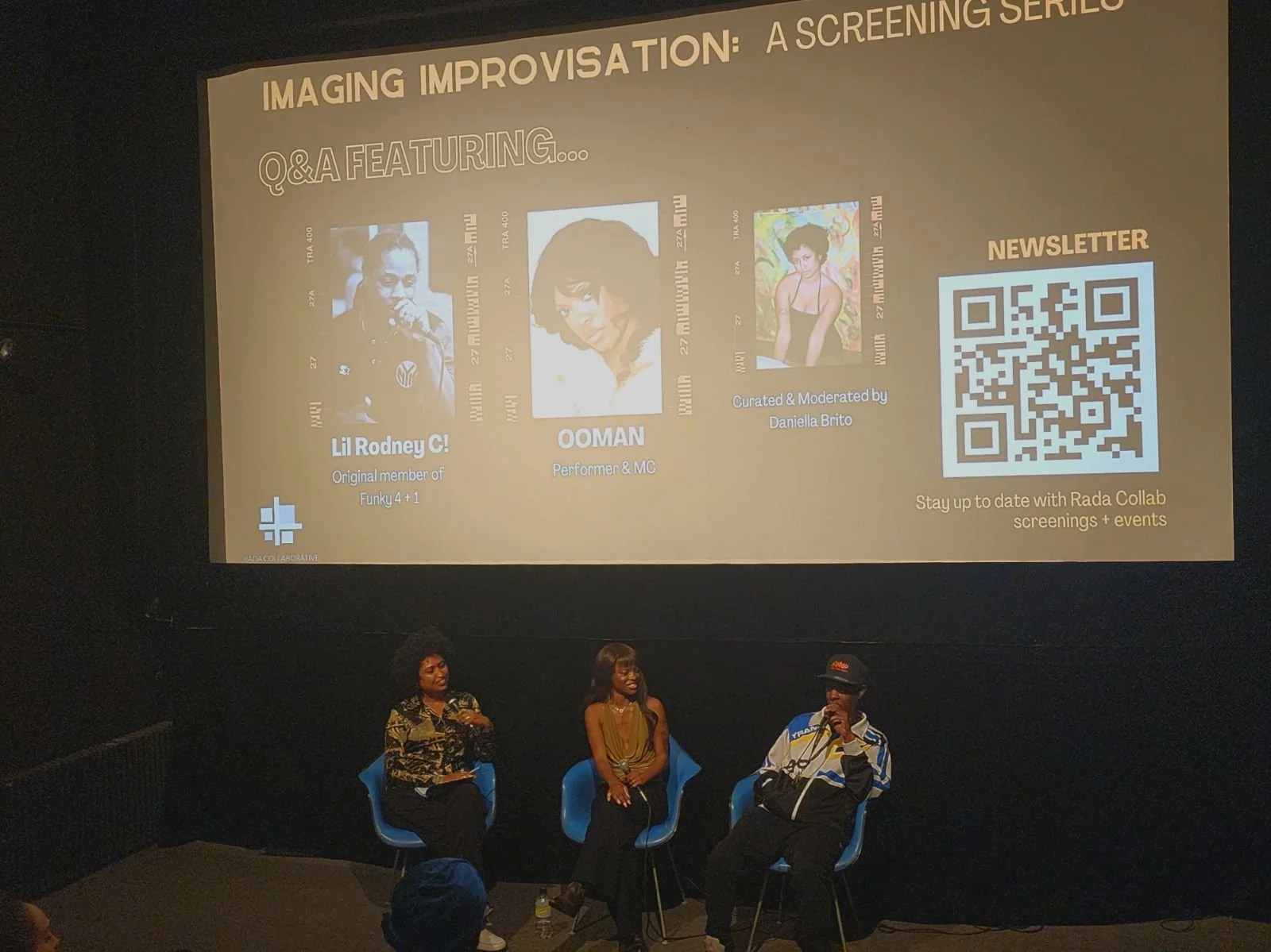 Panel discussion with three people seated on stage in front of a large screen displaying event information about a screening series called "Imaging Improvisation." The screen shows images and names of Lil Rodney C!, Ooman, and Daniela Brito, along with a QR code for newsletter subscription. The stage has a black background, and the panelists are seated in blue chairs.