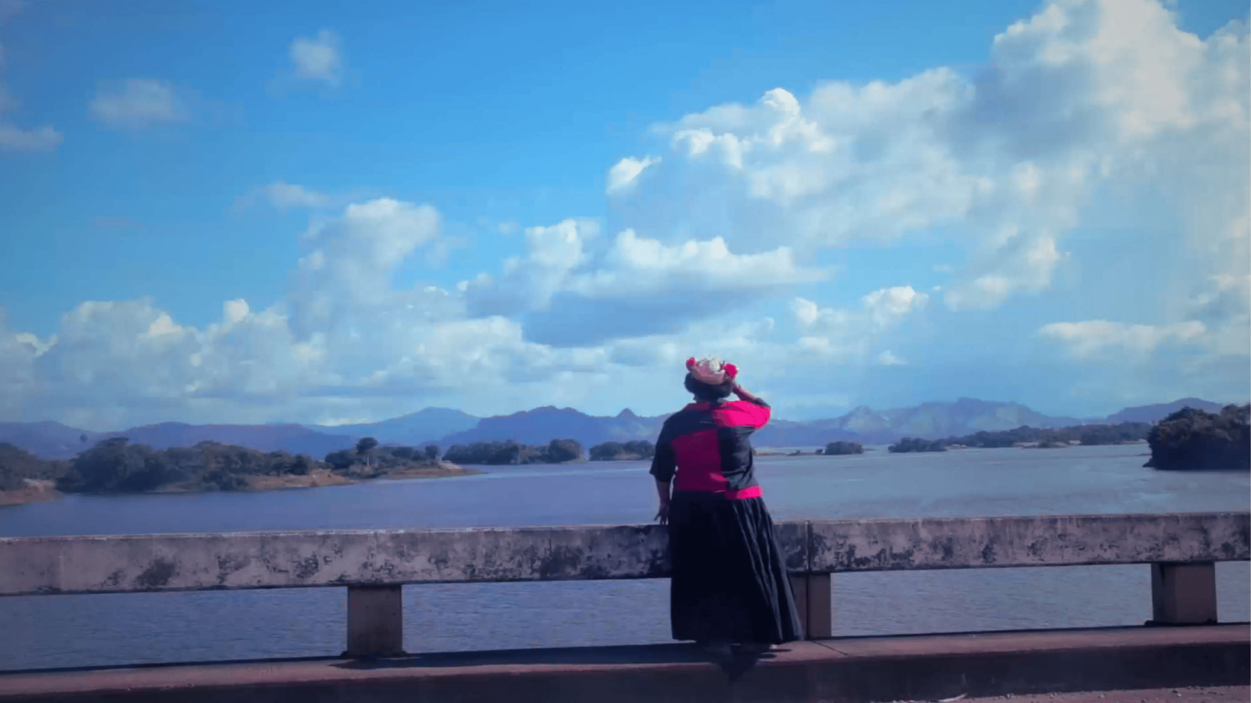 A woman in traditional dress in Panama and hat standing by a river looking at the water and distant mountains under a partly cloudy sky.