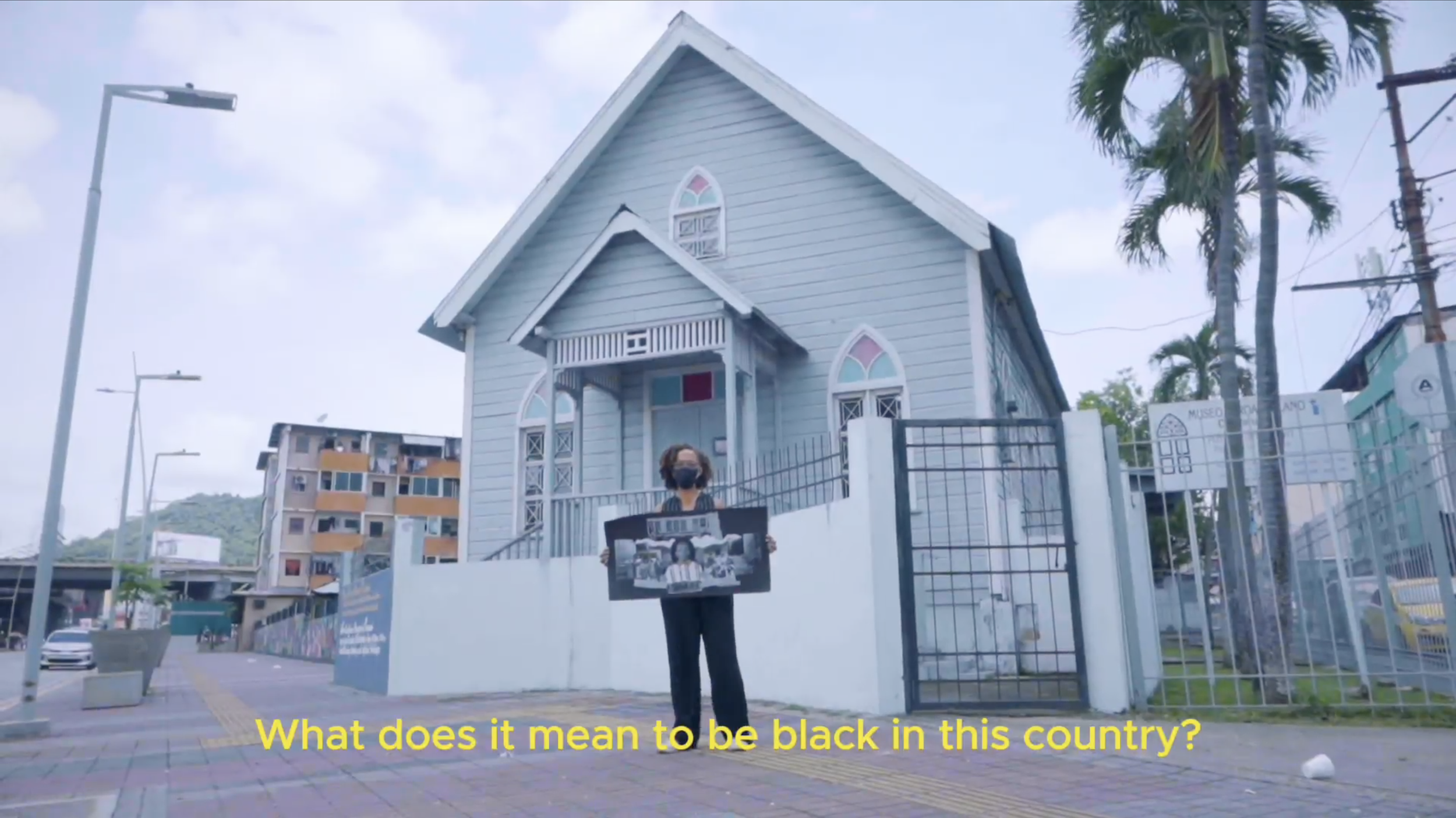A person standing outside a light blue house in Panama, holding a sign with a collage of images, wearing a black mask, with yellow subtitles that read, 'What does it mean to be black in this country?'