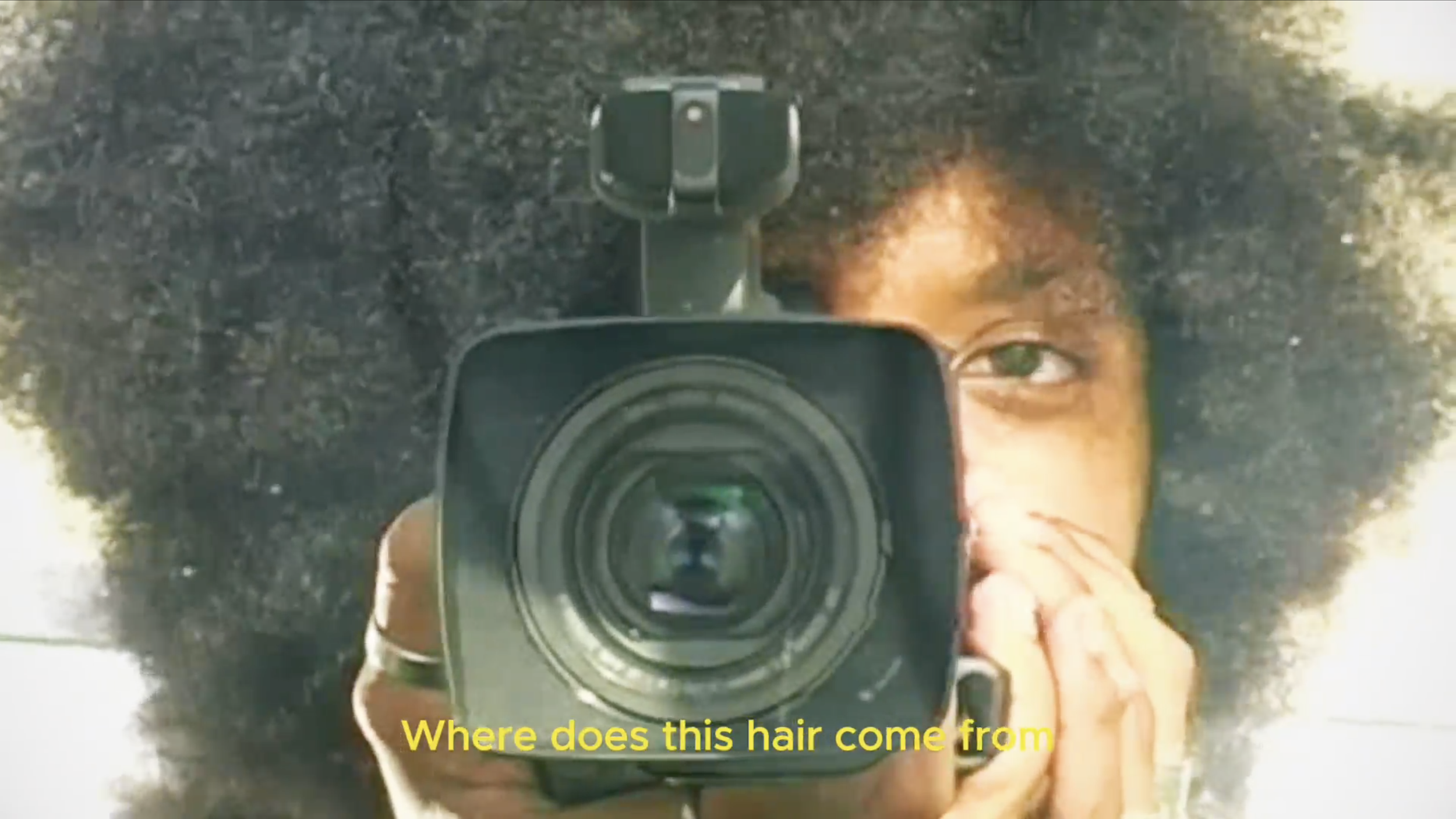 A Panamanian woman or person with curly hair holding a camera, taking a selfie in a mirror, with yellow text overlay reading 'Where does this hair come from'.