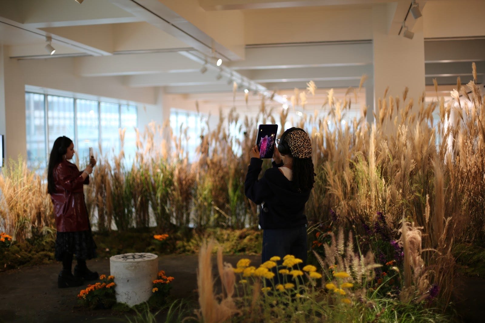 Two women are in an indoor garden with tall dried plants and flowers, one is taking a photo with her phone, and the other is posing for the picture.