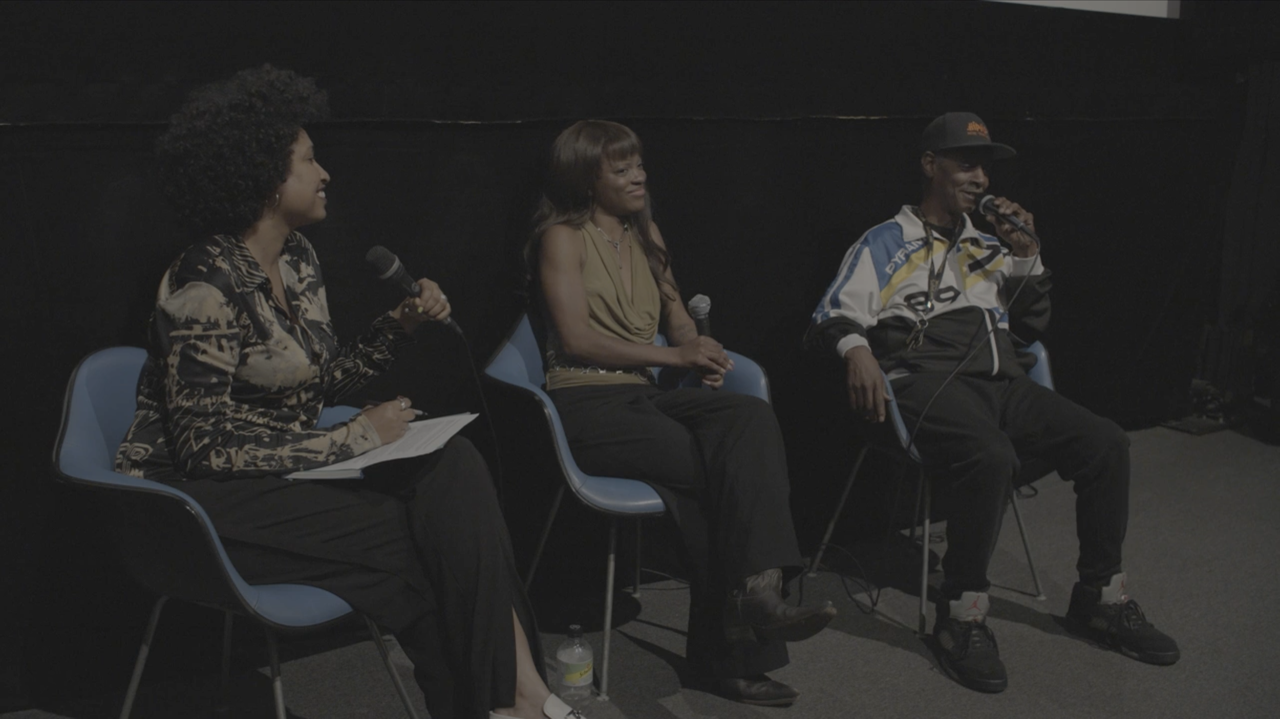 Three people sit on blue chairs on a stage, participating in a panel discussion. The person on the left is a woman holding a microphone and taking notes. The center is a woman with long hair, smiling and holding a microphone. The person on the right is a man wearing a baseball cap, a jacket, and sneakers, speaking into a microphone.