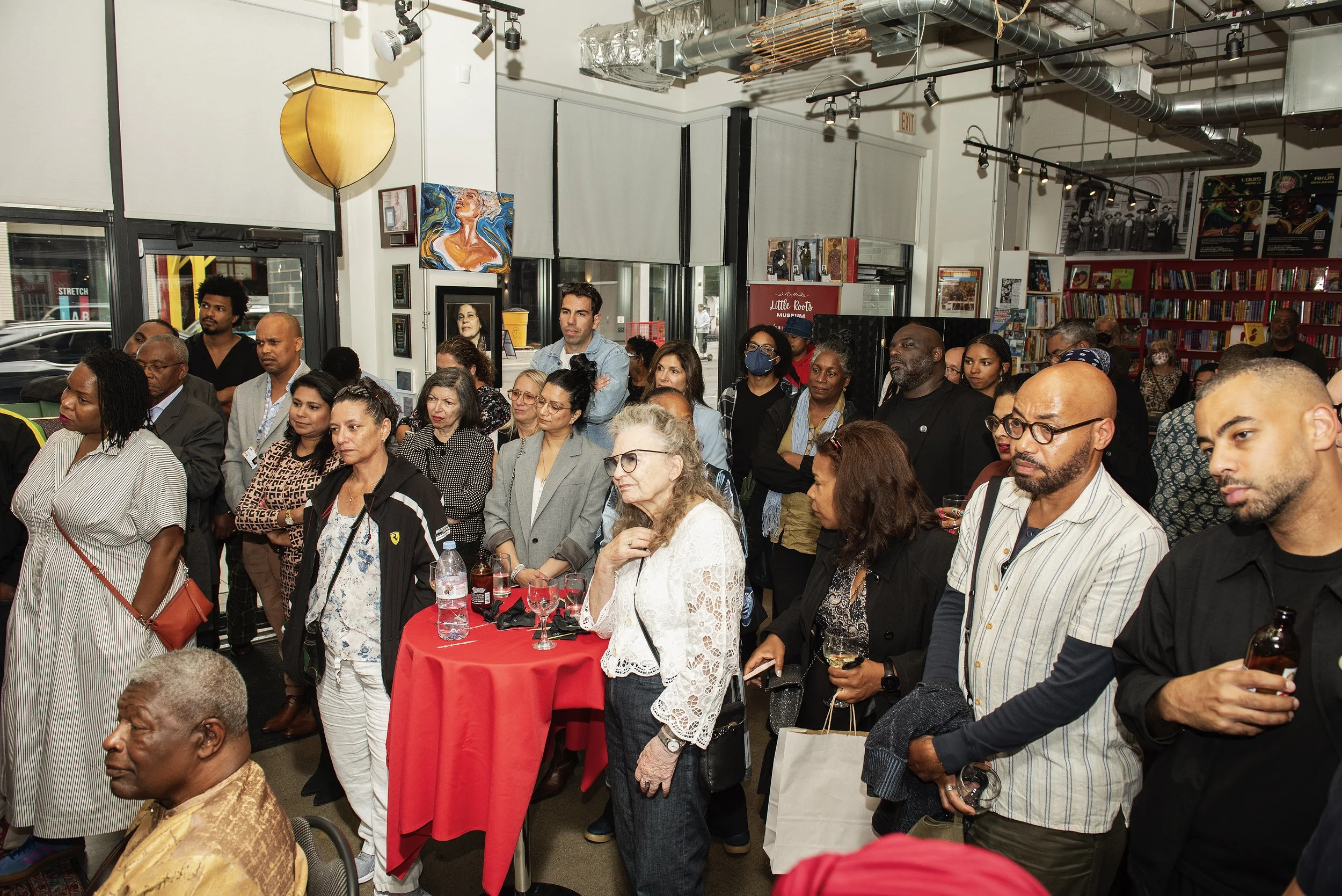 A diverse group of people attending an event at a bookstore or gallery, some standing around tables with drinks, inside a space decorated with artwork and books.