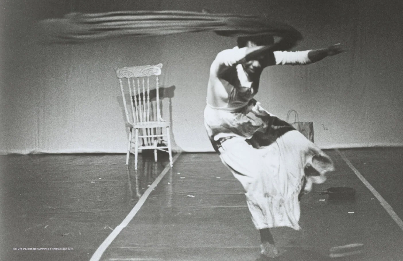 A woman dancing in a room with a chair and a bag in the background. She is wearing a long skirt and a top, captured mid-movement with her arms outstretched and her head tilted.