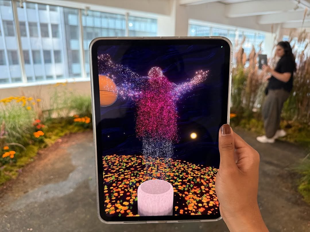 A person holding a tablet with a digital hologram of a human figure made of pink and purple particles, standing above a circular pink platform, with a spacecraft and a starry sky in the background.