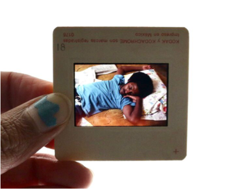 A person holding a compact digital camera with an LCD screen displaying a photo of a young girl lying down on a bed, petting a small animal.
