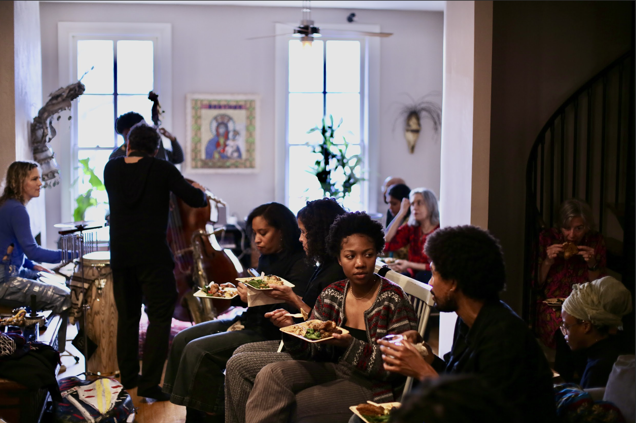 People seated and eating food in a cozy living room with large windows, a painting on the wall, and musical instruments being played by musicians.