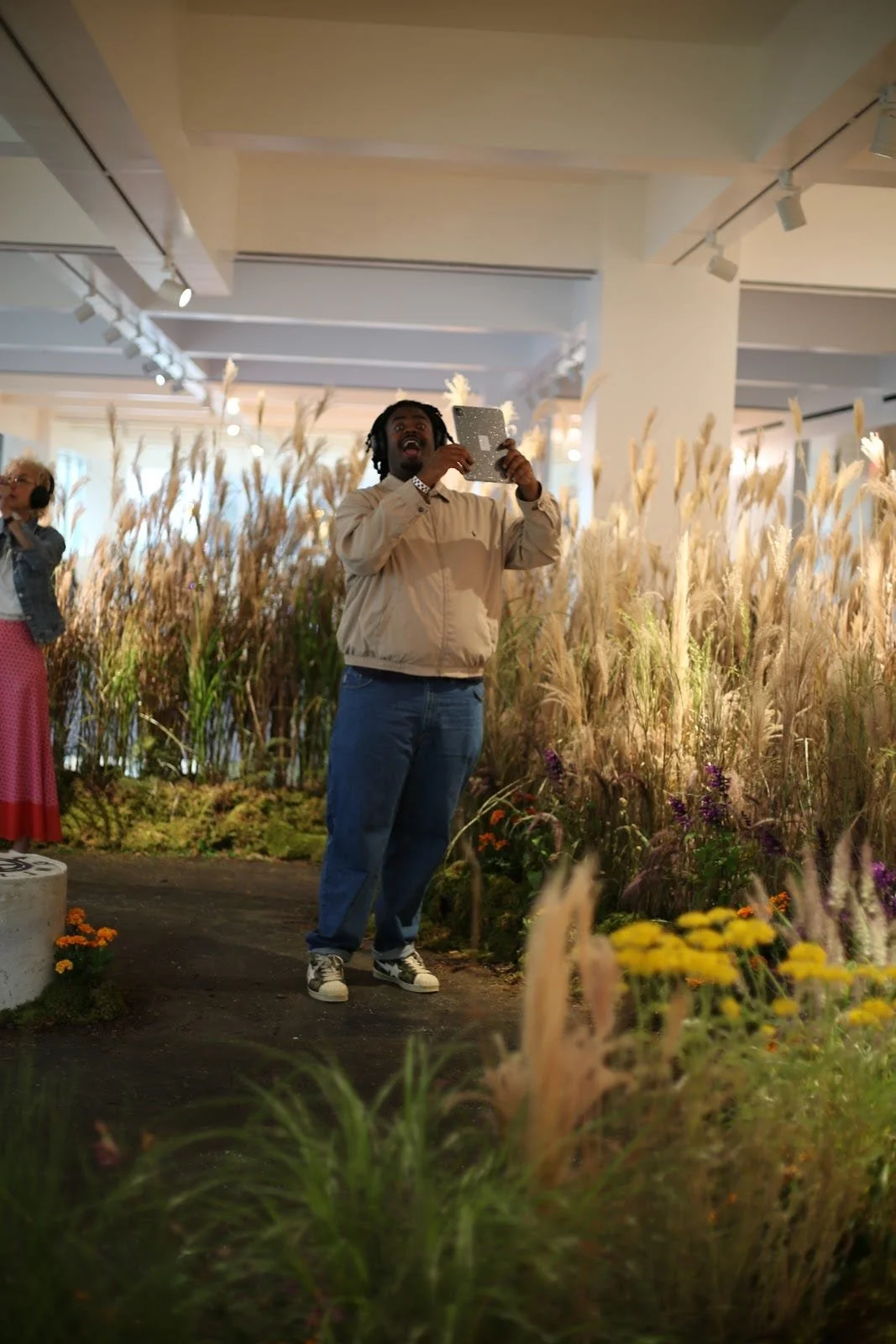 A man taking a photo with his phone in an indoor garden exhibit with tall grasses and flowers.