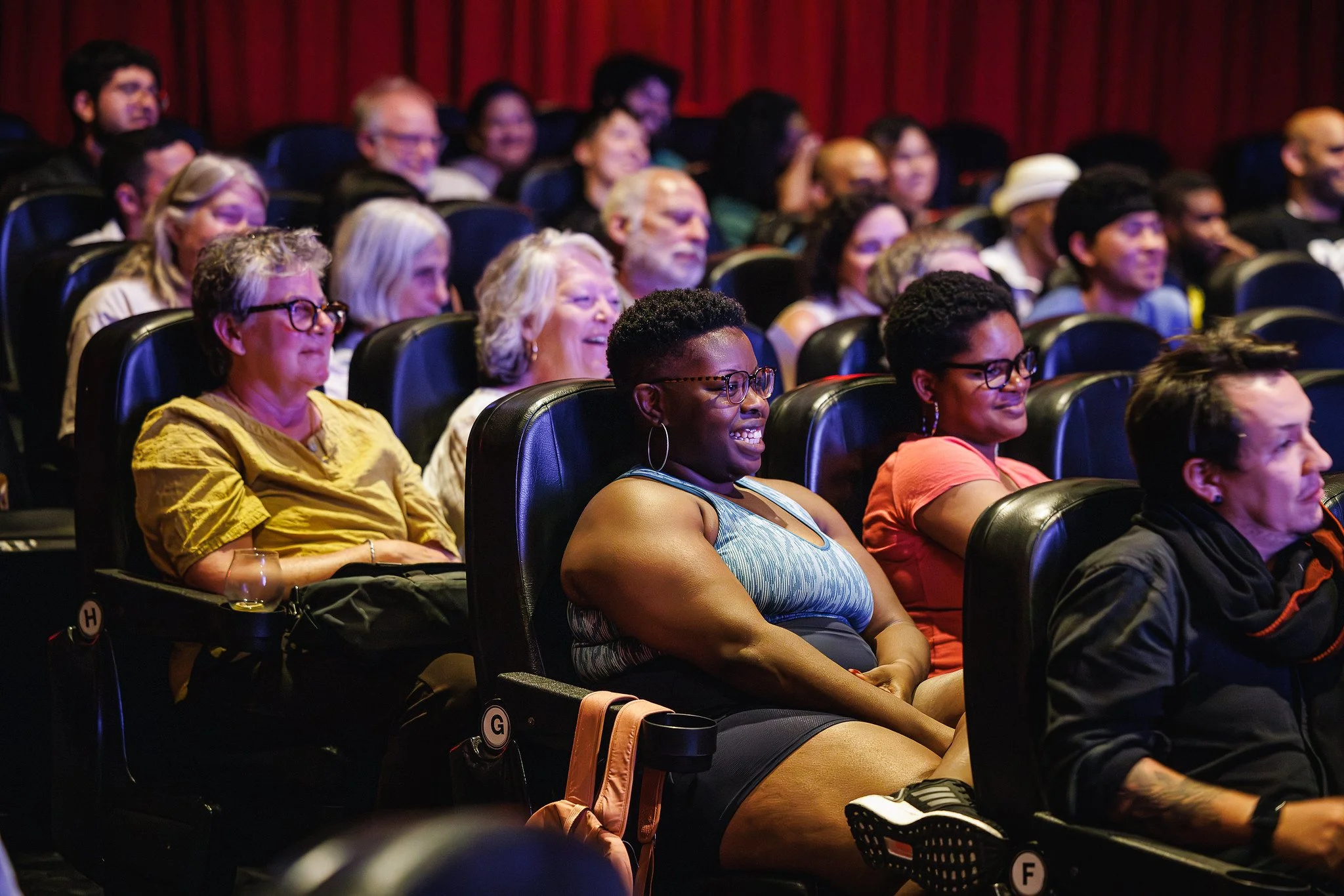 Audience members seated in a theater watching a performance, with some smiling and enjoying the event.