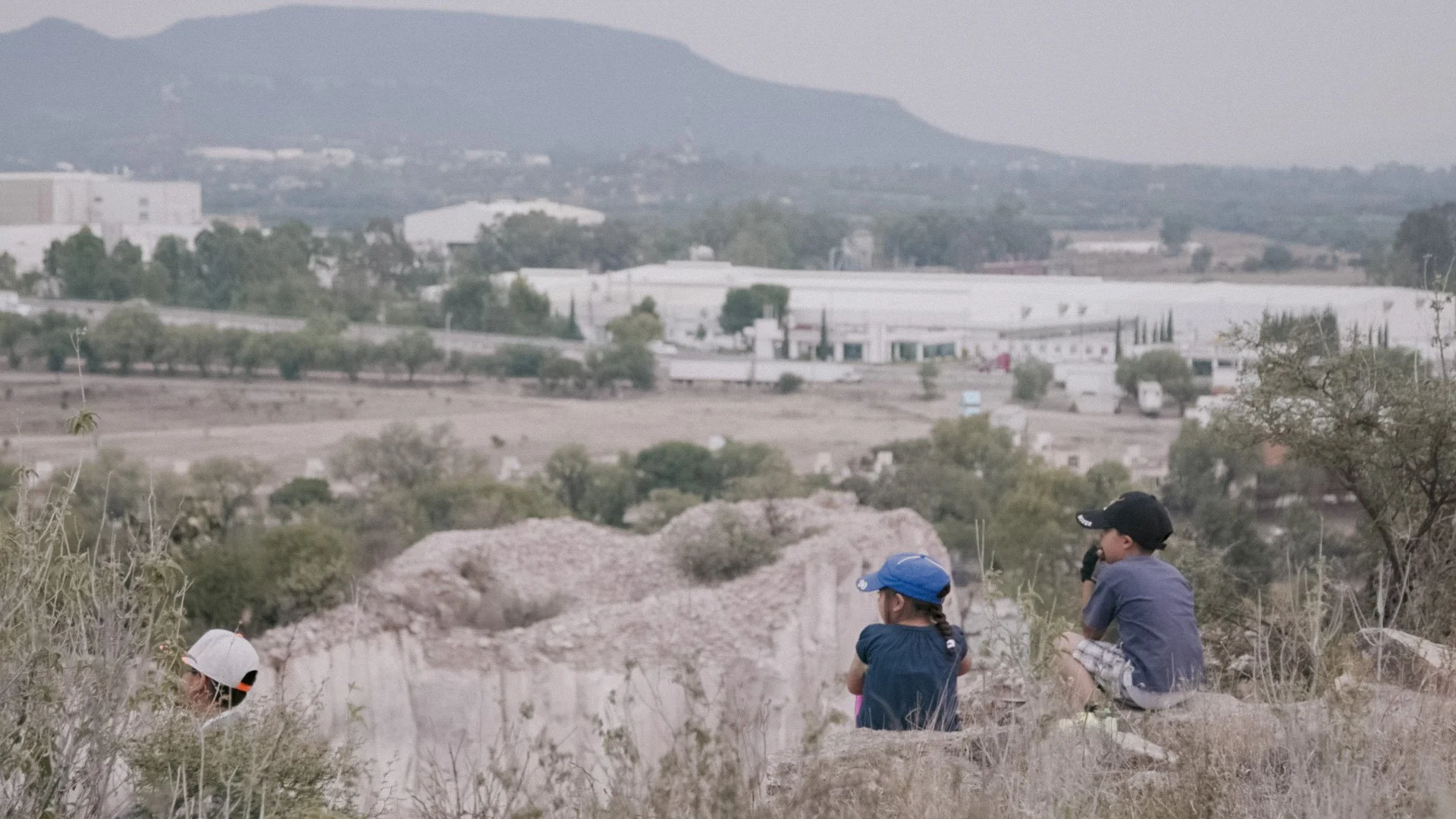 Three children sitting outdoors on a rocky hillside, looking into the distance, with a landscape of trees, industrial buildings, and mountains in the background.
