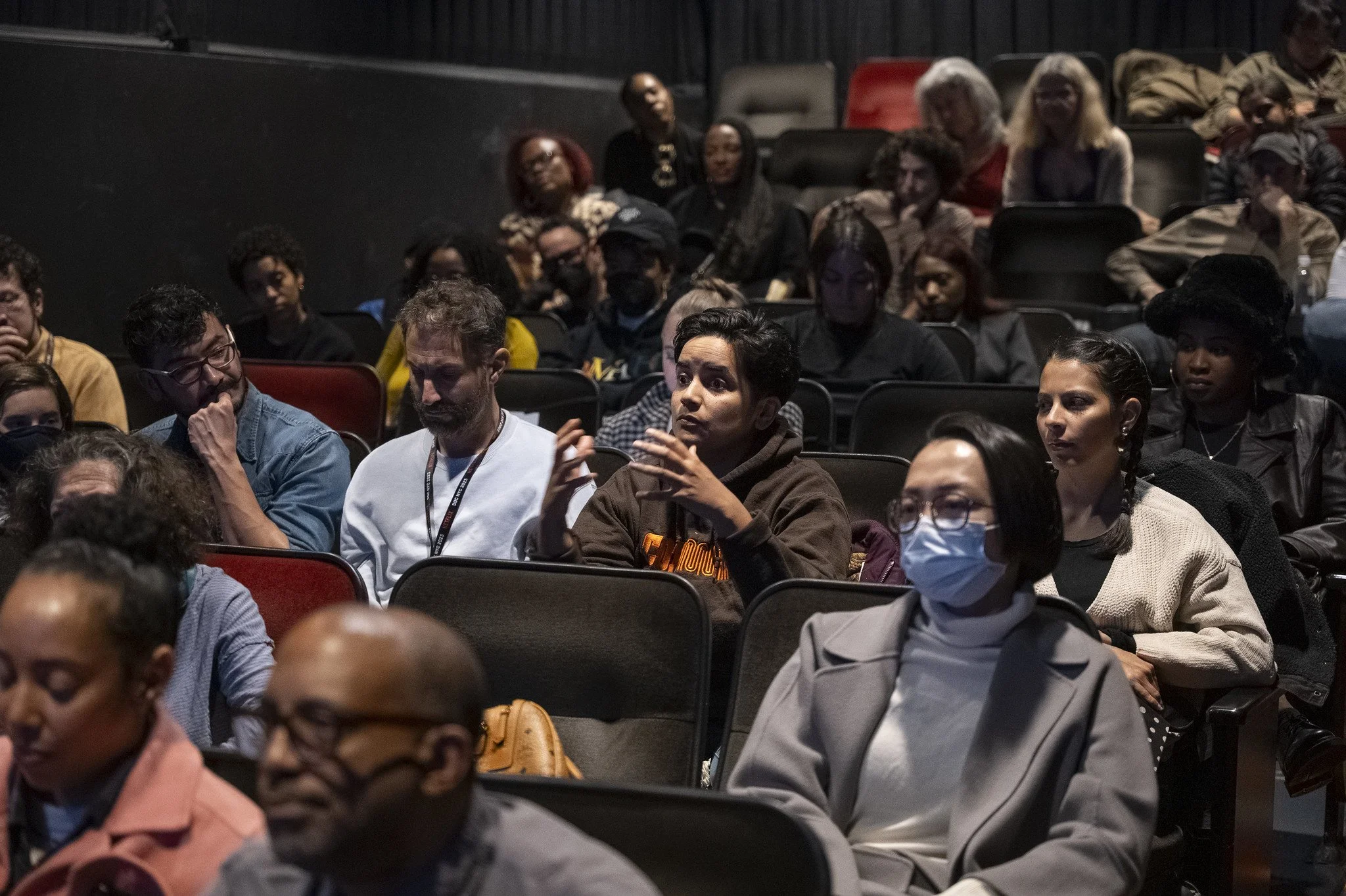Audience members sitting in a dark auditorium, some wearing face masks, listening attentively during a presentation or discussion.