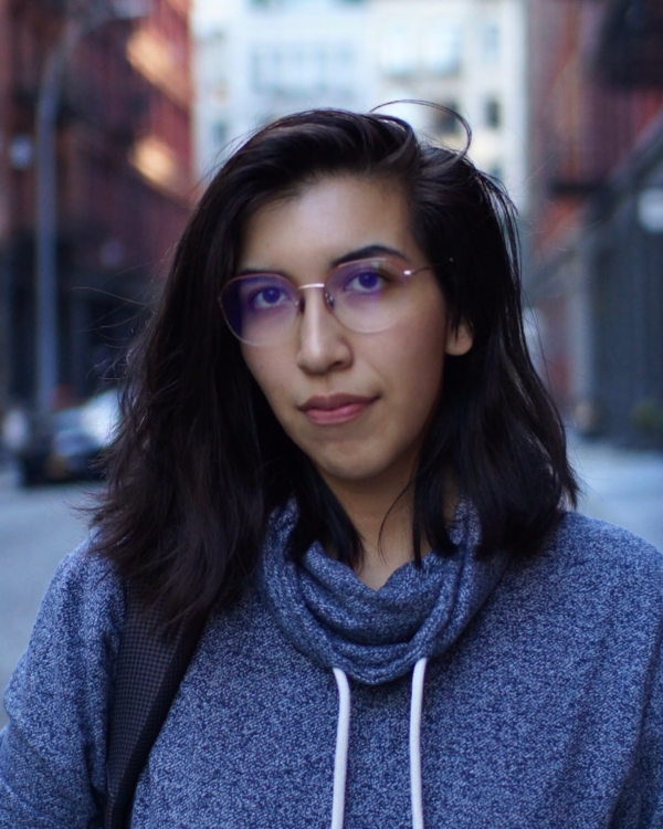 A young woman standing outdoors on a city street with buildings and cars in the background. She has shoulder-length dark hair, wears glasses with purple-tinted lenses, and is dressed in a blue hoodie with a matching scarf.