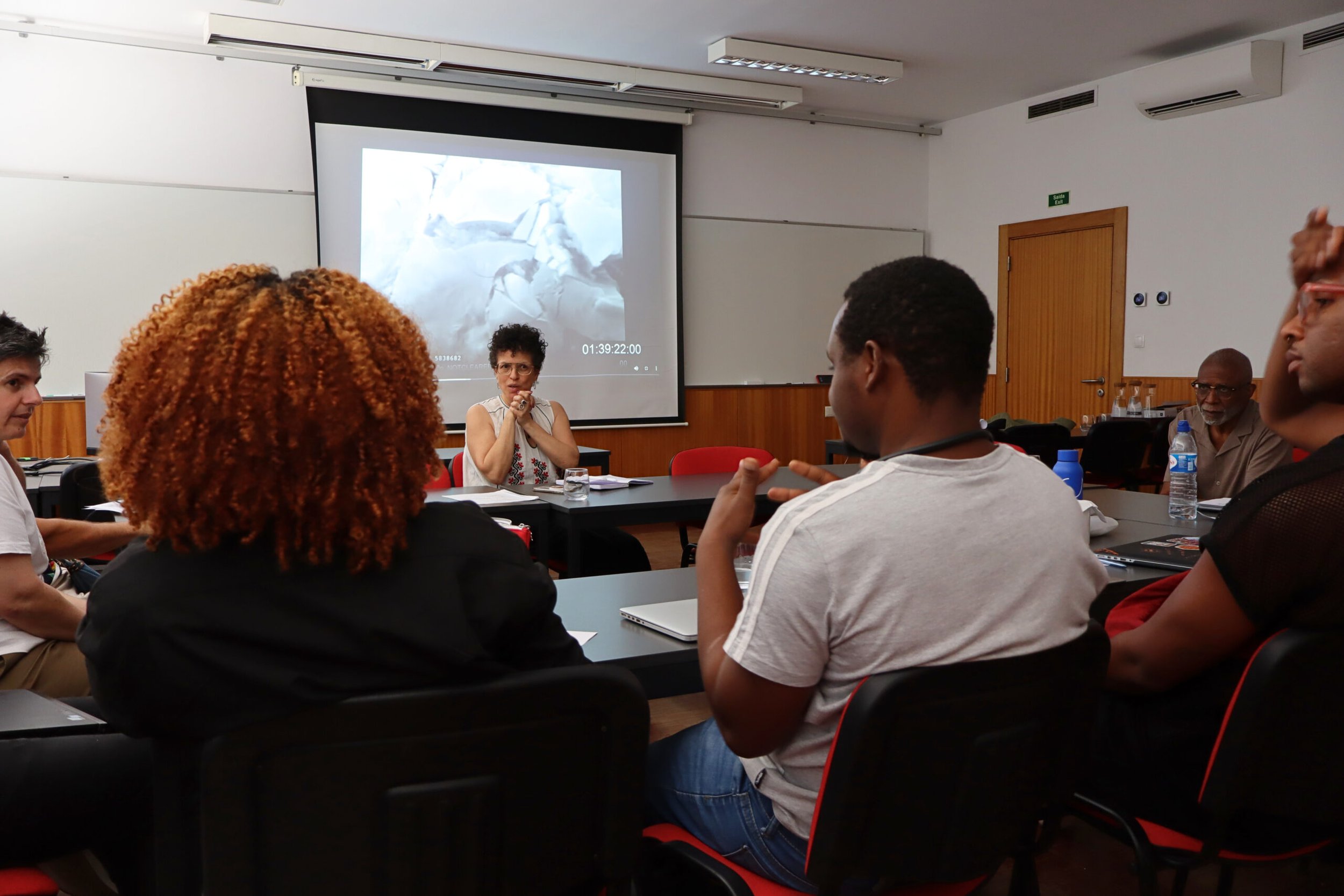 A woman is presenting to a group of diverse people in a classroom or conference room, with a large screen behind her displaying an image and a timestamp.