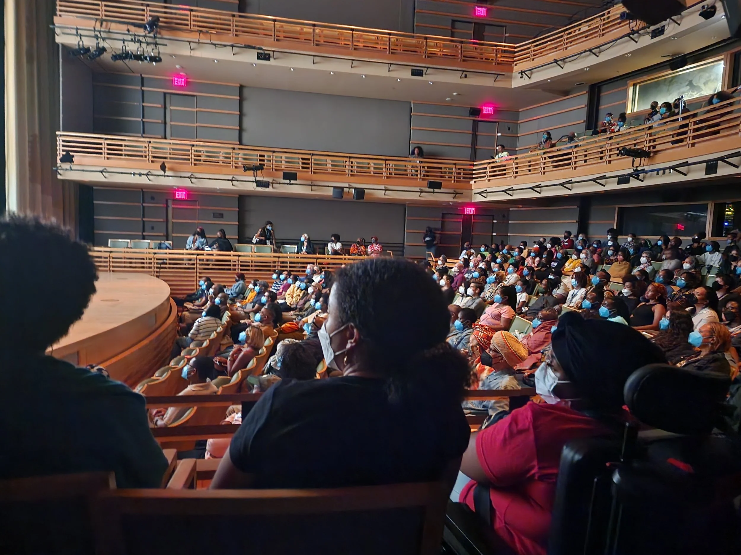 An audience seated in a theater watching a presentation, with some people on the upper balcony, all wearing face masks.
