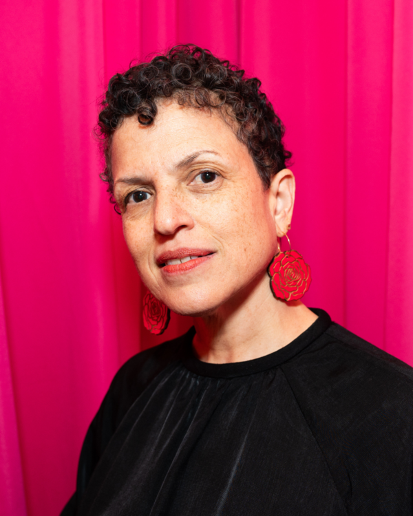 A woman with short, curly hair wearing red floral earrings and a black top stands in front of a pink curtain.