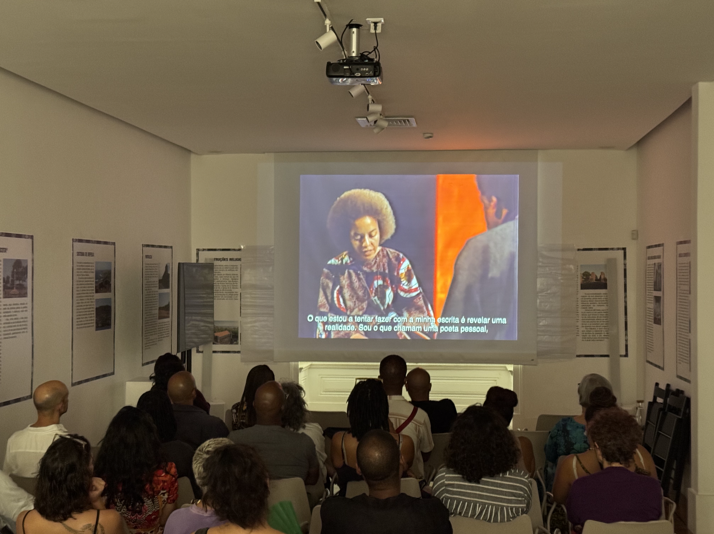 People seated in an exhibition room watching a video presentation on a large screen, with posters on the walls.