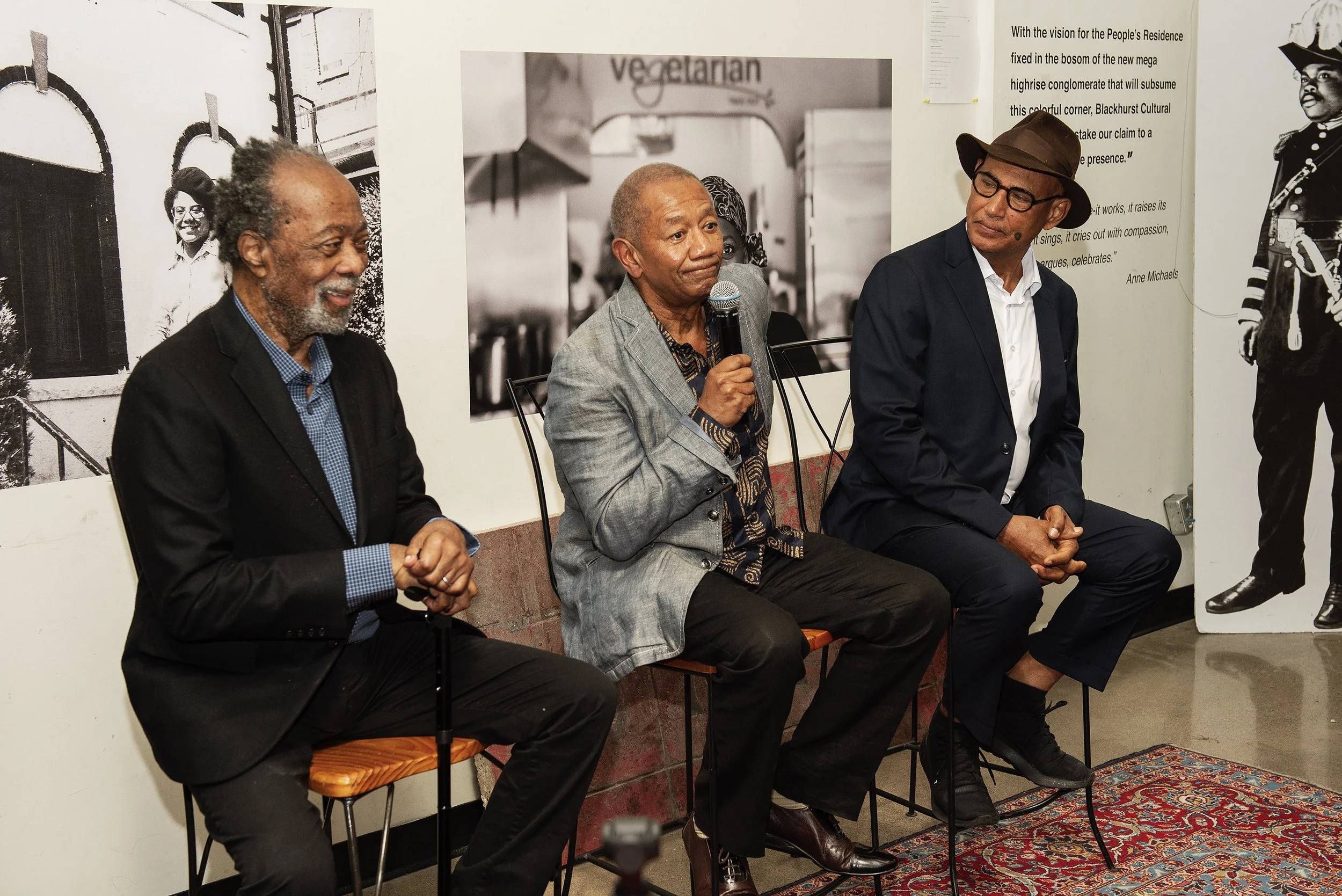 Three men sitting on chairs in a room, with posters and photos on the wall behind them. The man in the middle is speaking into a microphone. The floor has a rug.