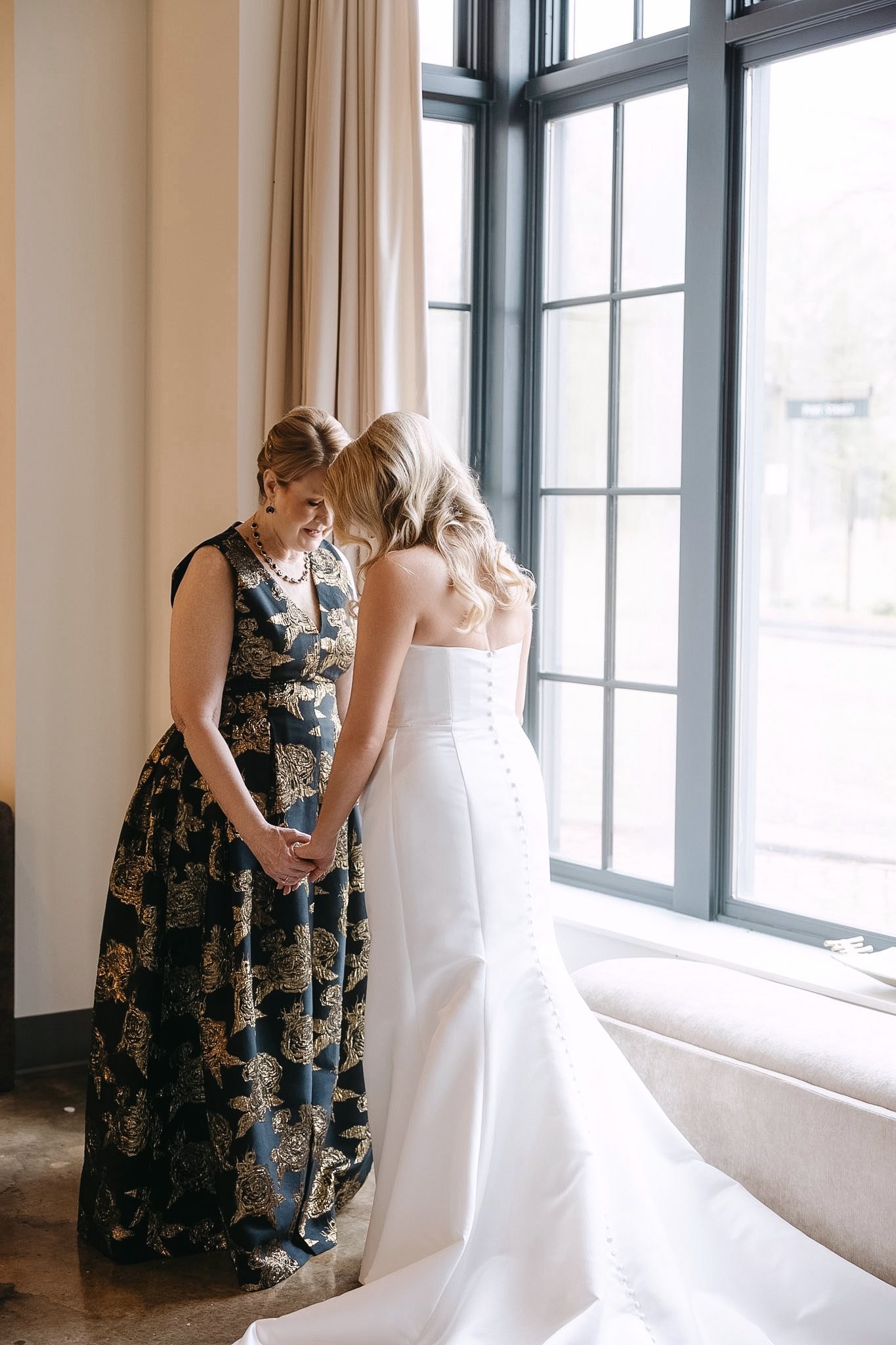 Mother-and-daughter moment at The Velveteen wedding venue in Madison, MS, holding hands forehead-to-forehead beside a softly curtained window on the wedding day