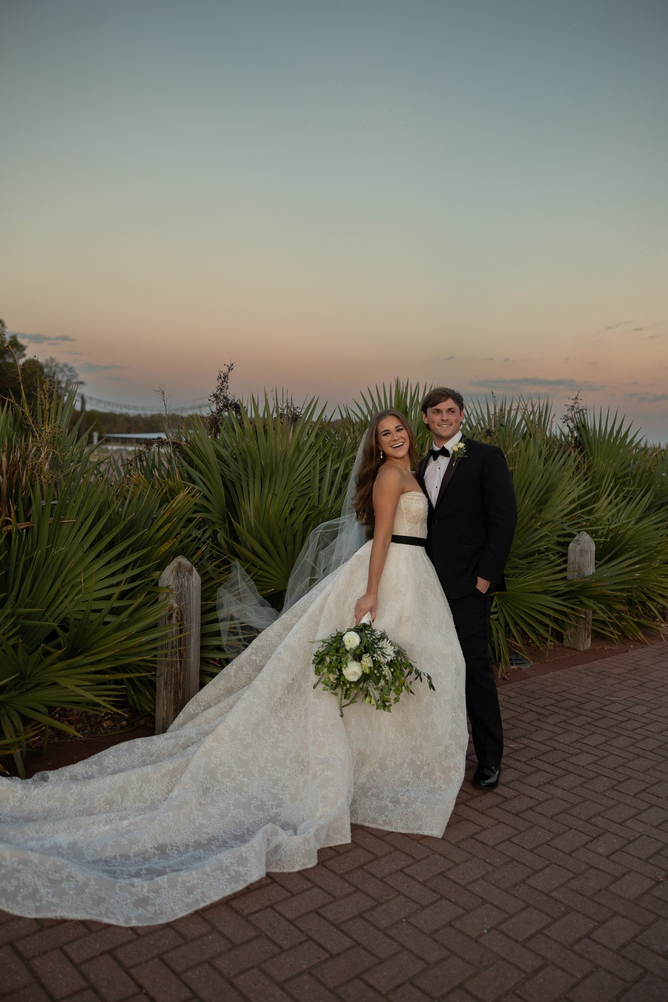 Bride and groom at sunset at The Velveteen wedding venue in Madison, MS, the bride in a lace gown with a white bouquet beside her groom in a black tuxedo