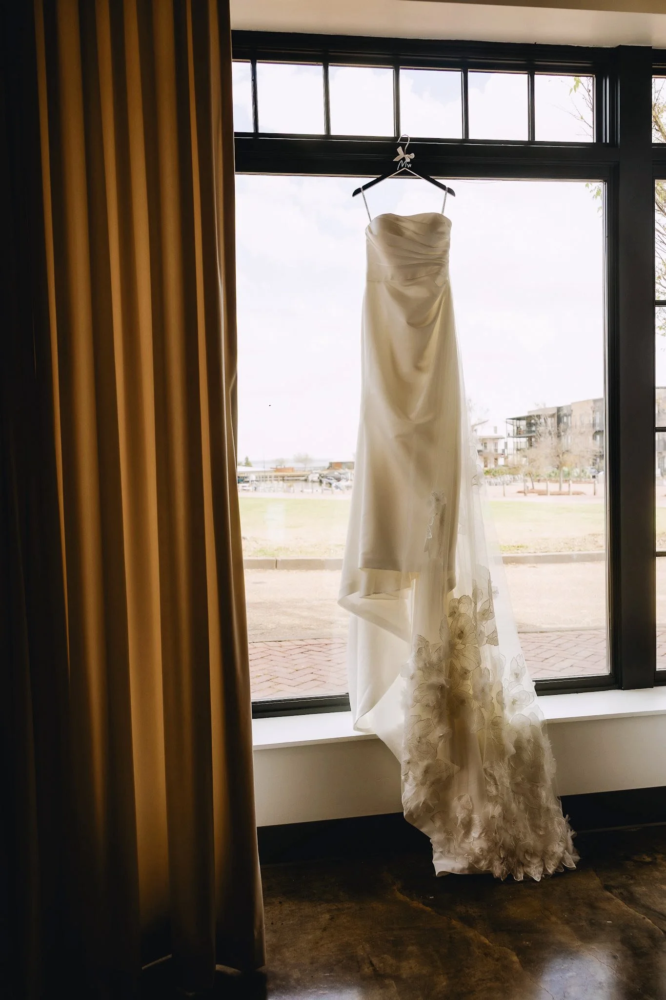 Wedding gown hung in the bridal suite at The Velveteen wedding venue in Madison, MS, framed by a soft curtained window with views beyond the glass