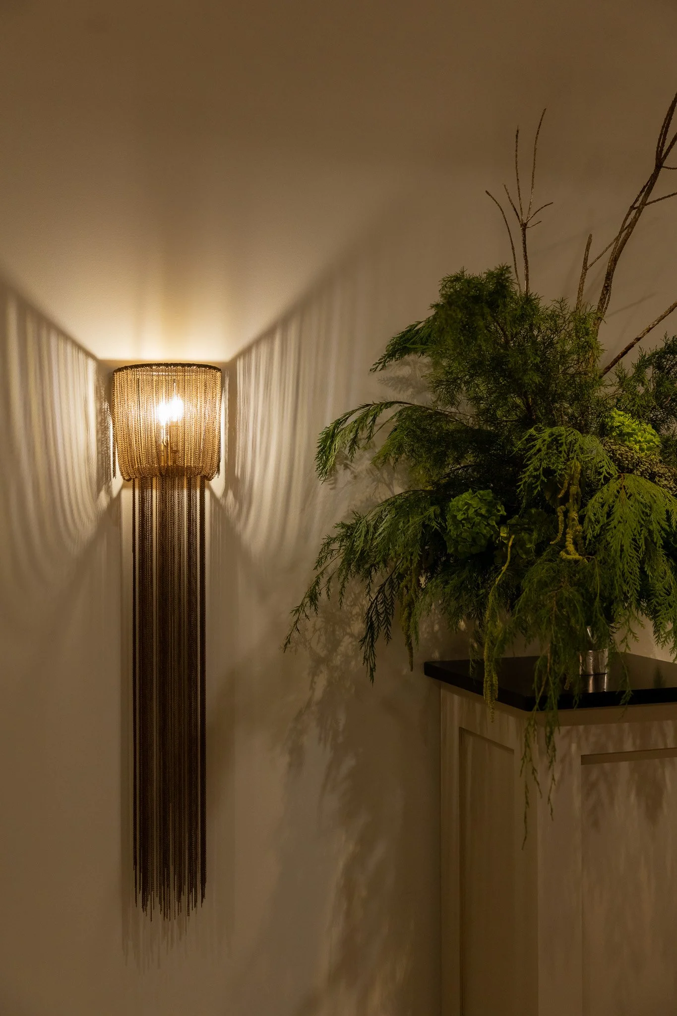 Detail at The Velveteen in Madison, MS, with a tall woven wall sconce casting warm light beside a leafy green plant on a dark cabinet