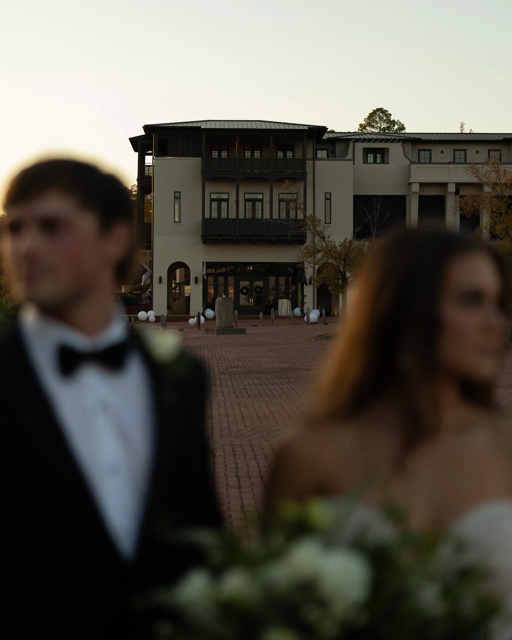 Outdoor sunset gathering at The Velveteen wedding venue in Madison, MS, with guests in tuxedos mingling in front of the illuminated building