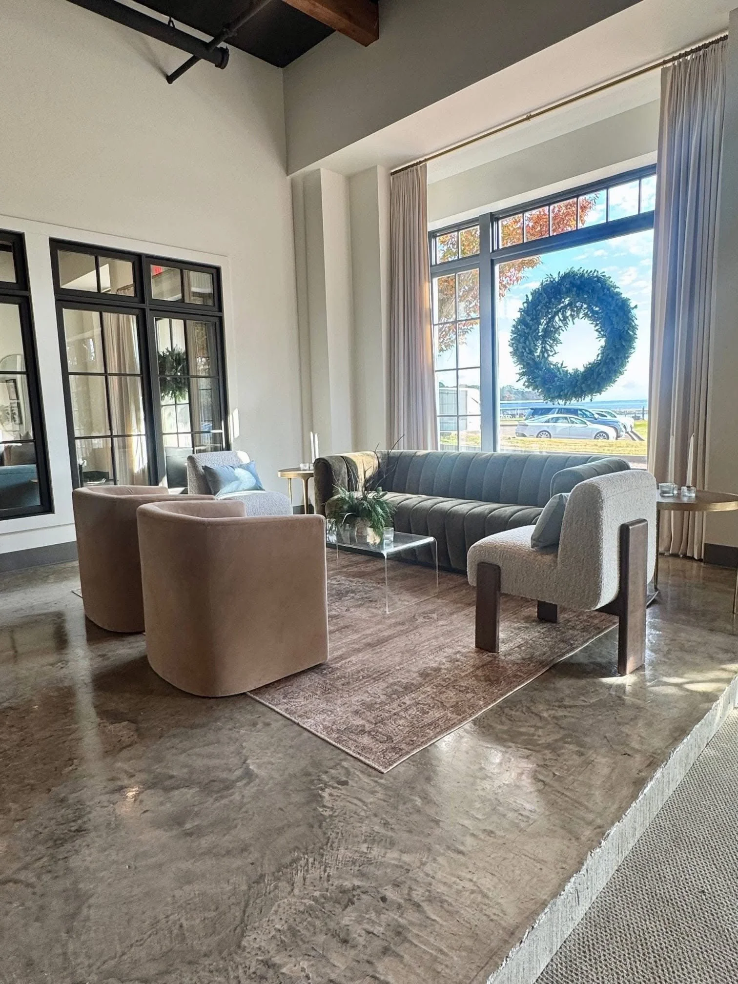 Lounge area at The Velveteen wedding venue in Madison, MS, with beige armchairs, a charcoal sofa, and a glass coffee table positioned by tall windows overlooking the water