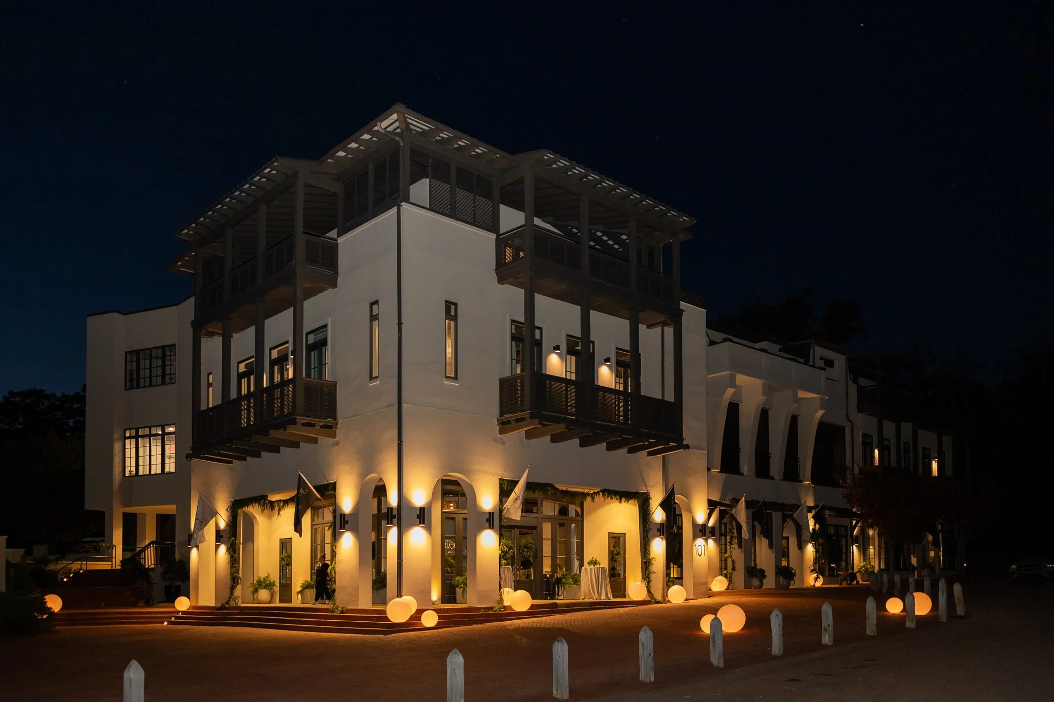 Evening exterior of The Velveteen wedding venue in Madison, MS, glowing with warm uplighting along the multi-story facade, balconies, and rooftop terrace