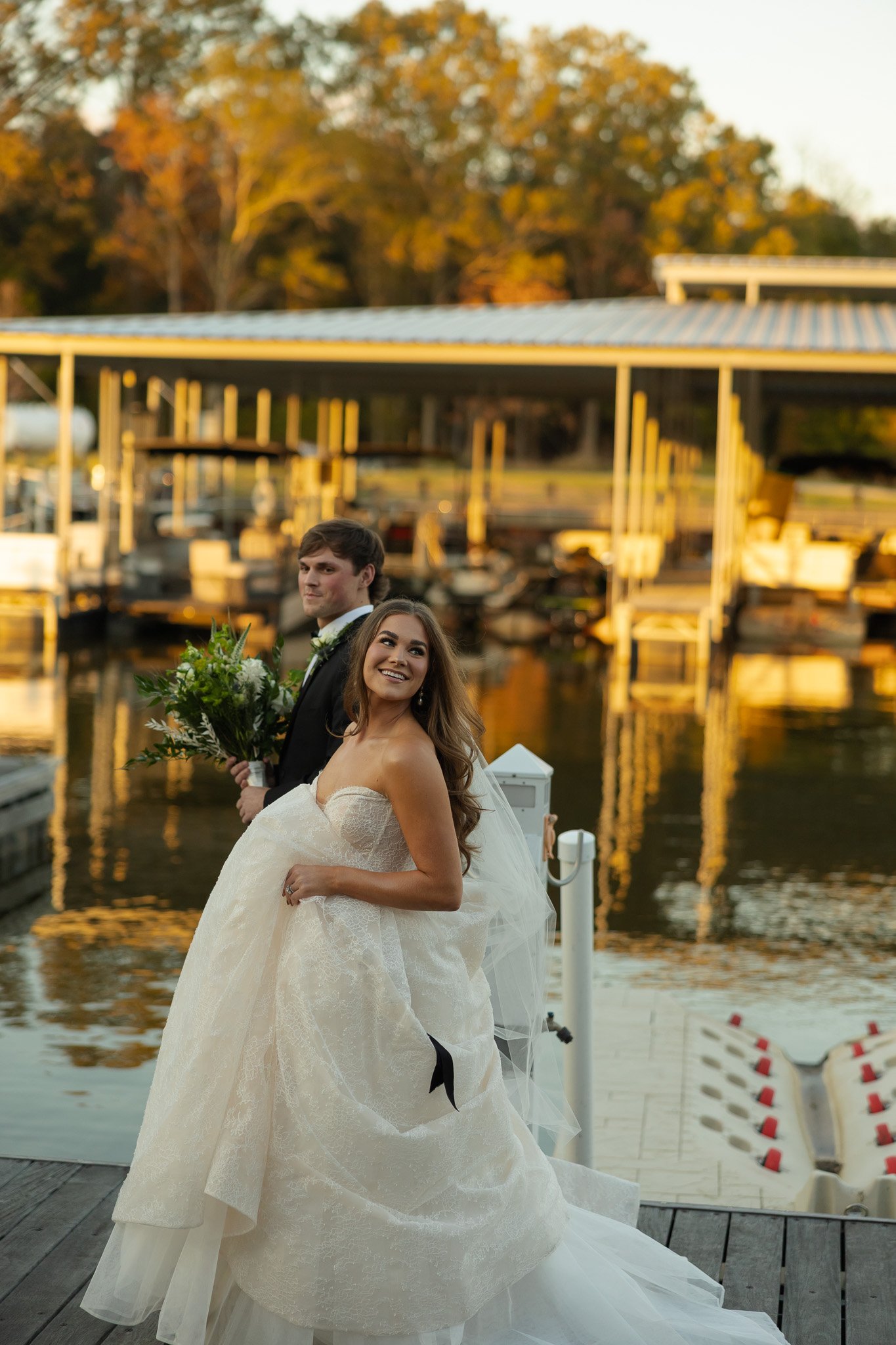 Wedding portrait on the dock at The Velveteen wedding venue in Madison, MS, the couple standing by the marina with autumn trees behind them at sunset