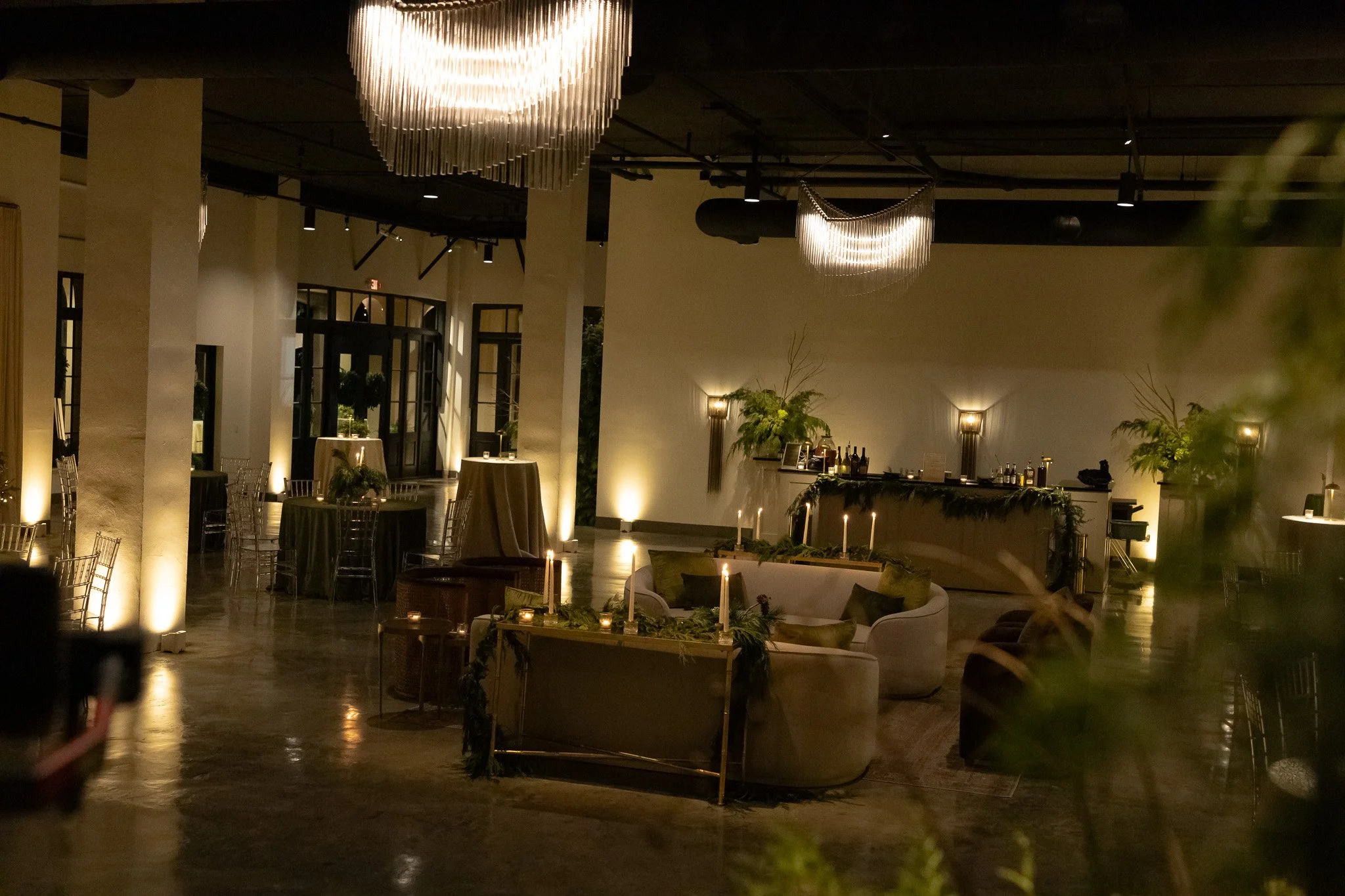 Evening reception at The Velveteen wedding venue in Madison, MS, with crystal chandeliers, candlelit round tables, a bar in back, and lush potted greenery