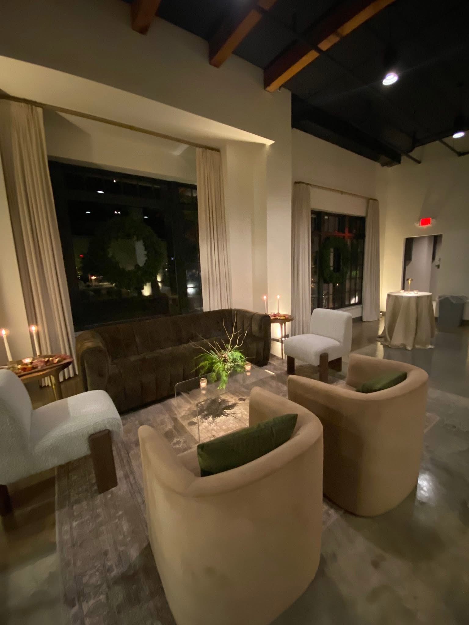 Reception lounge at The Velveteen wedding venue in Madison, MS, with beige armchairs, a brown leather sofa, lit pillar candles, and a wedding cake table beneath exposed black ceiling beams