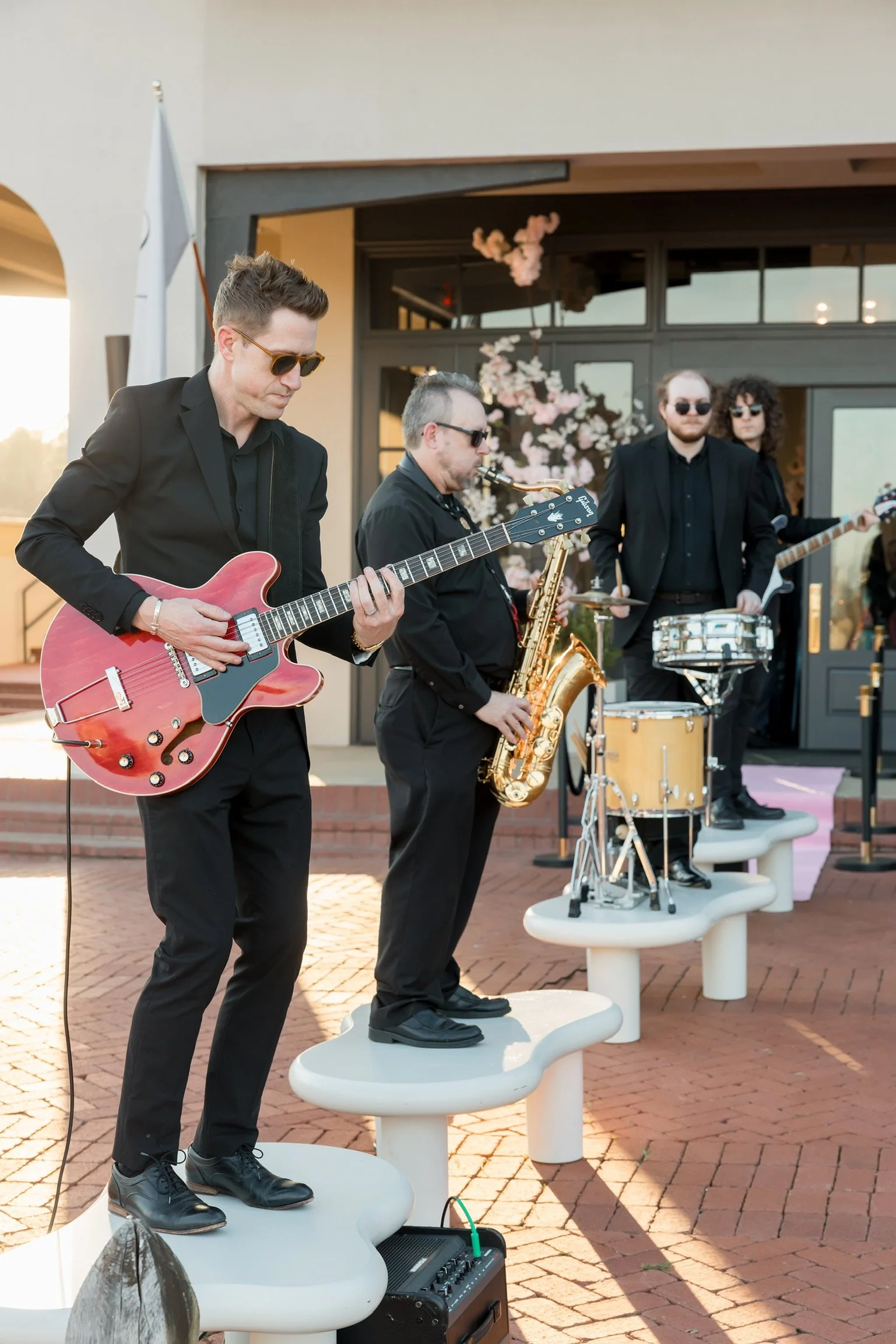 Live band performance at The Velveteen wedding venue in Madison, MS, with musicians in black suits playing guitar, saxophone, drums, and bass on a pink-carpeted patio at sunset