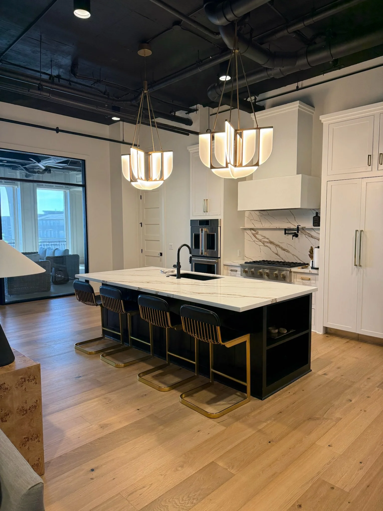 Modern kitchen at The Velveteen wedding venue in Madison, MS, with white cabinetry, marble backsplash, a black-and-gold island with stools, and an adjacent lounge through sliding glass doors