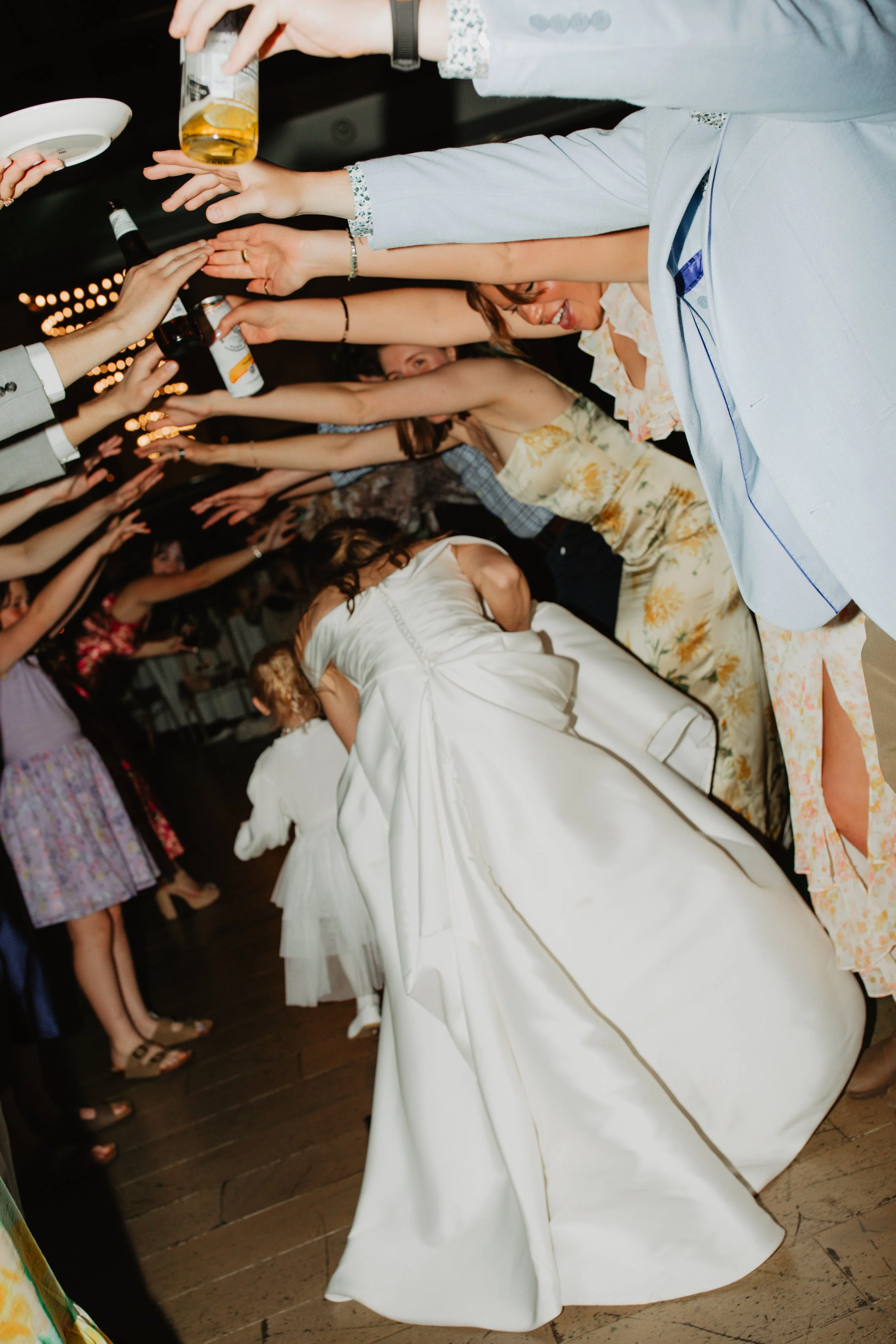 Women in floral dresses and the bride in a white wedding gown dancing together at a wedding reception, with guests holding drinks and reaching out their hands.