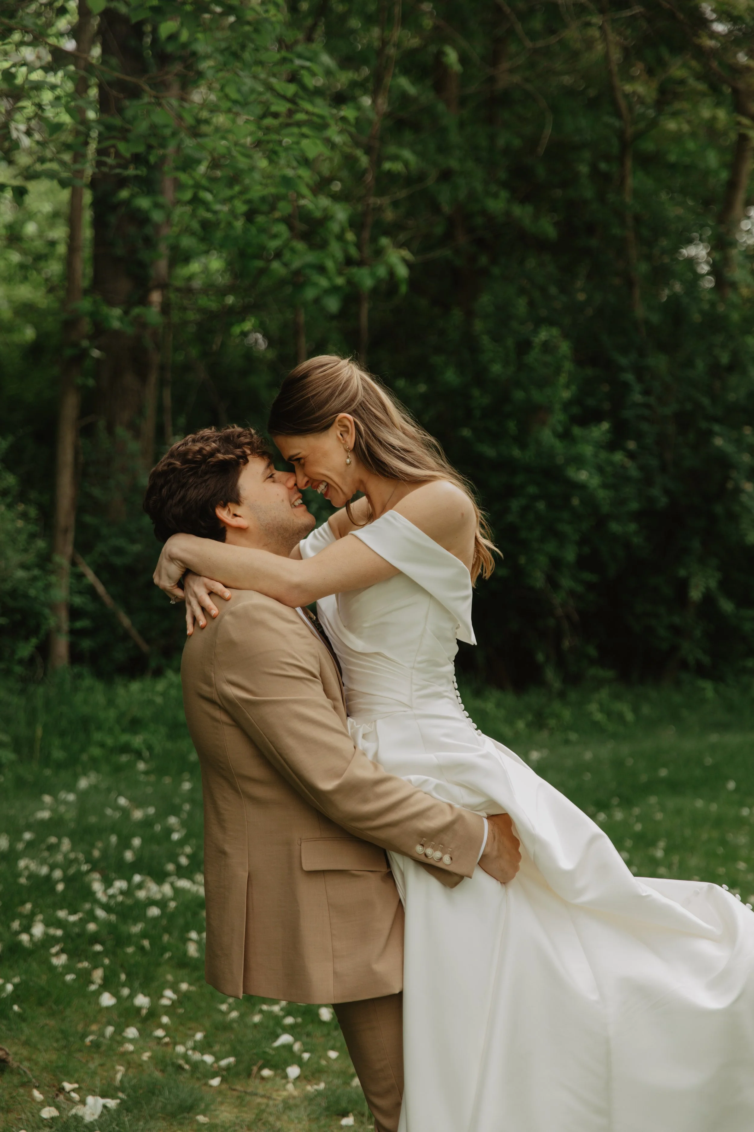 A newlywed couple in wedding attire embracing outdoors in a lush green setting.
