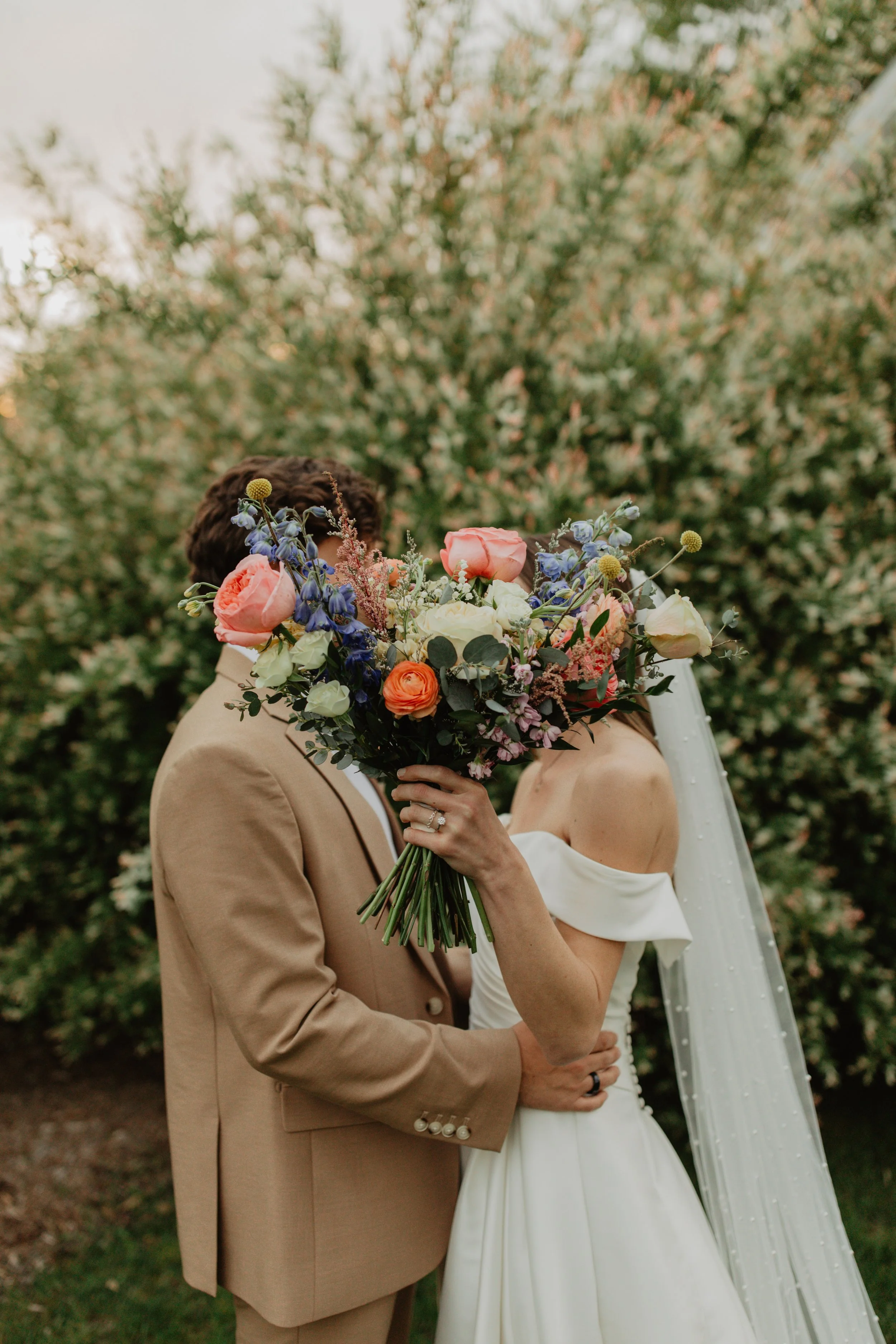 A bride and groom share a kiss behind a large bouquet of colorful flowers during their wedding outdoors.