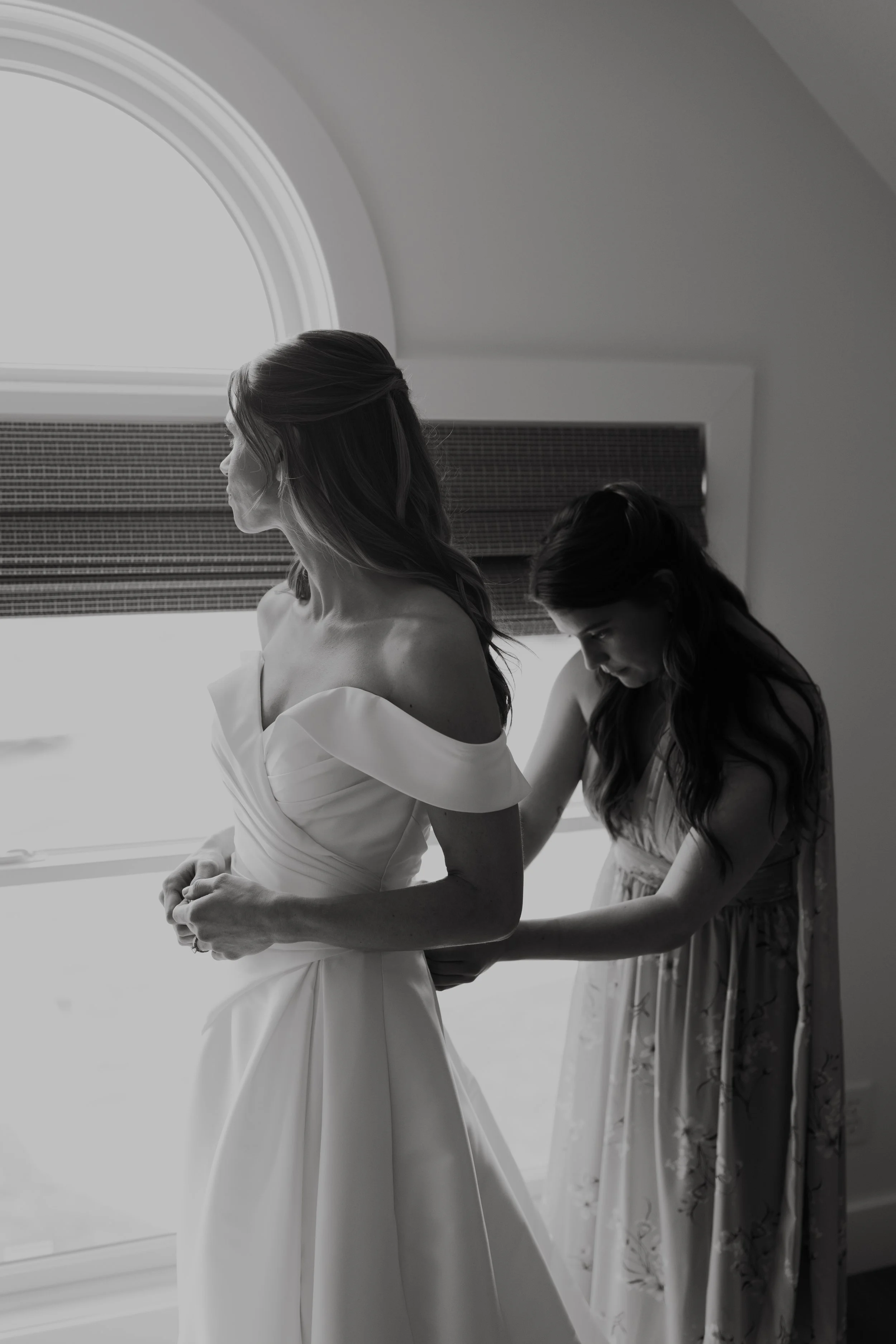 A bride in an off-shoulder wedding gown is being helped with her dress by a bridesmaid in a floral dress, as they stand near a window.