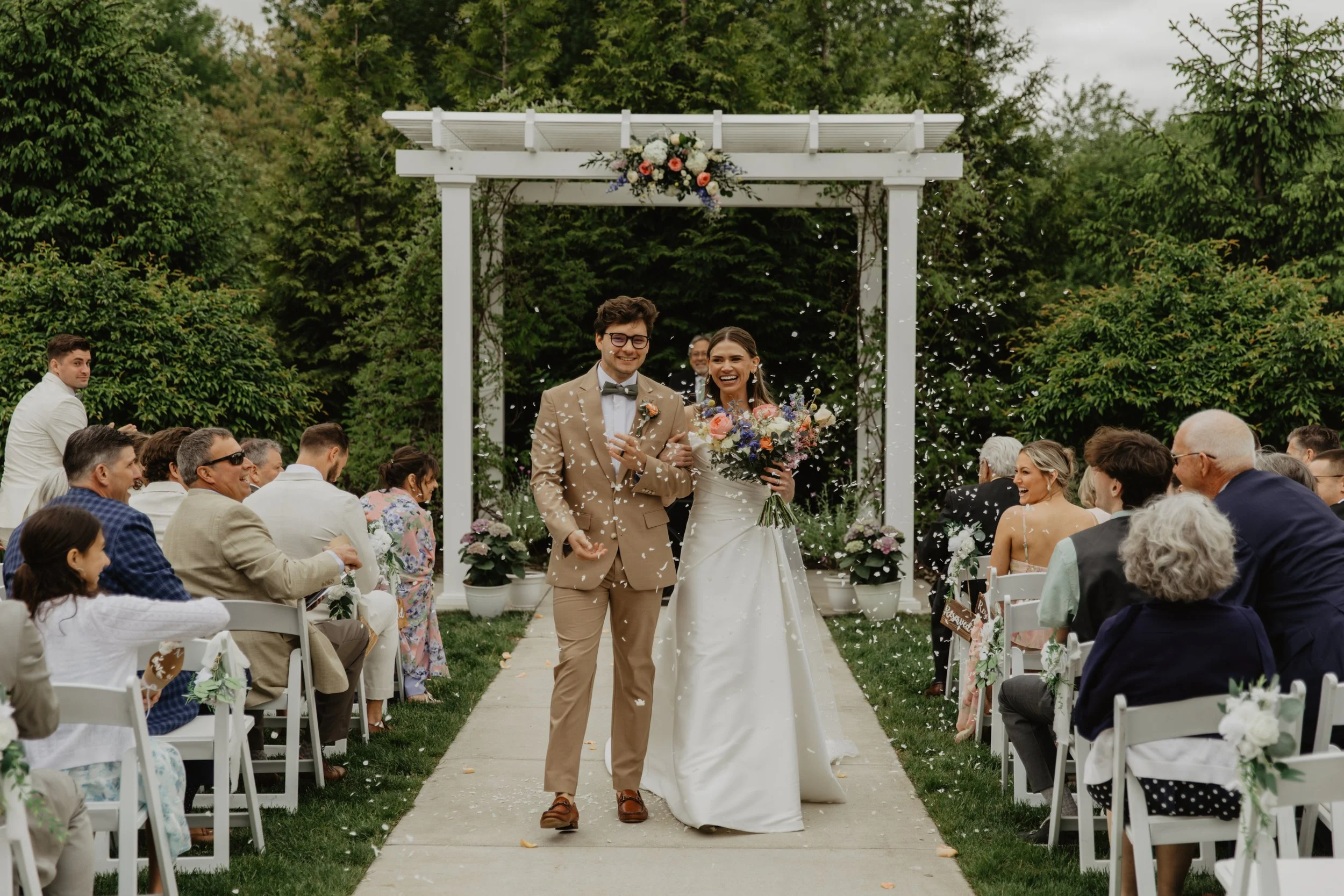A newlywed couple walking down the aisle at their outdoor wedding ceremony, surrounded by guests, with confetti falling, greenery, and a decorative arch with flowers.