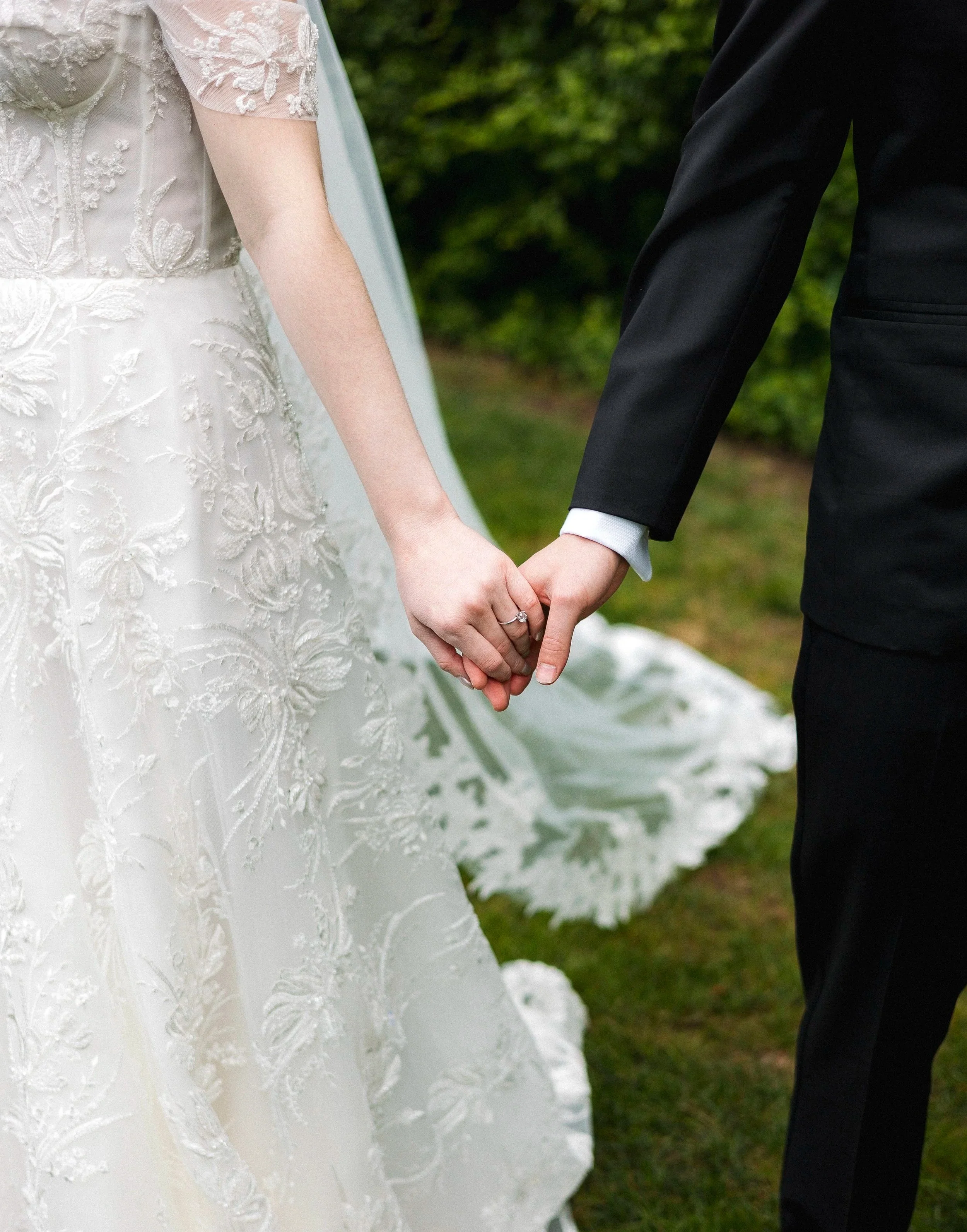 Close-up of a bride and groom holding hands during their wedding, with the bride wearing a lace wedding dress and the groom in a black suit.
