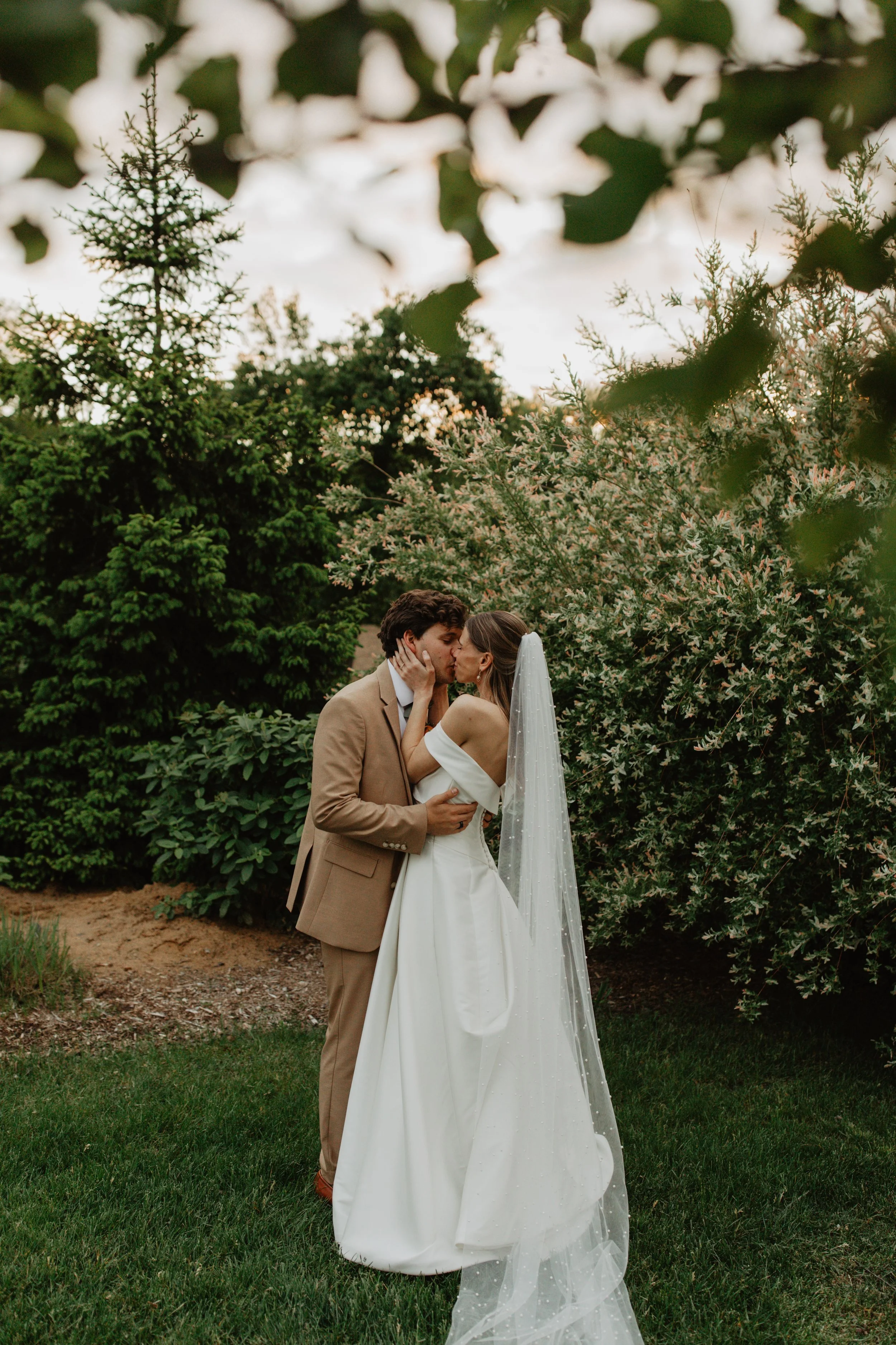 A bride and groom share a kiss outdoors, surrounded by greenery and trees, during their wedding.