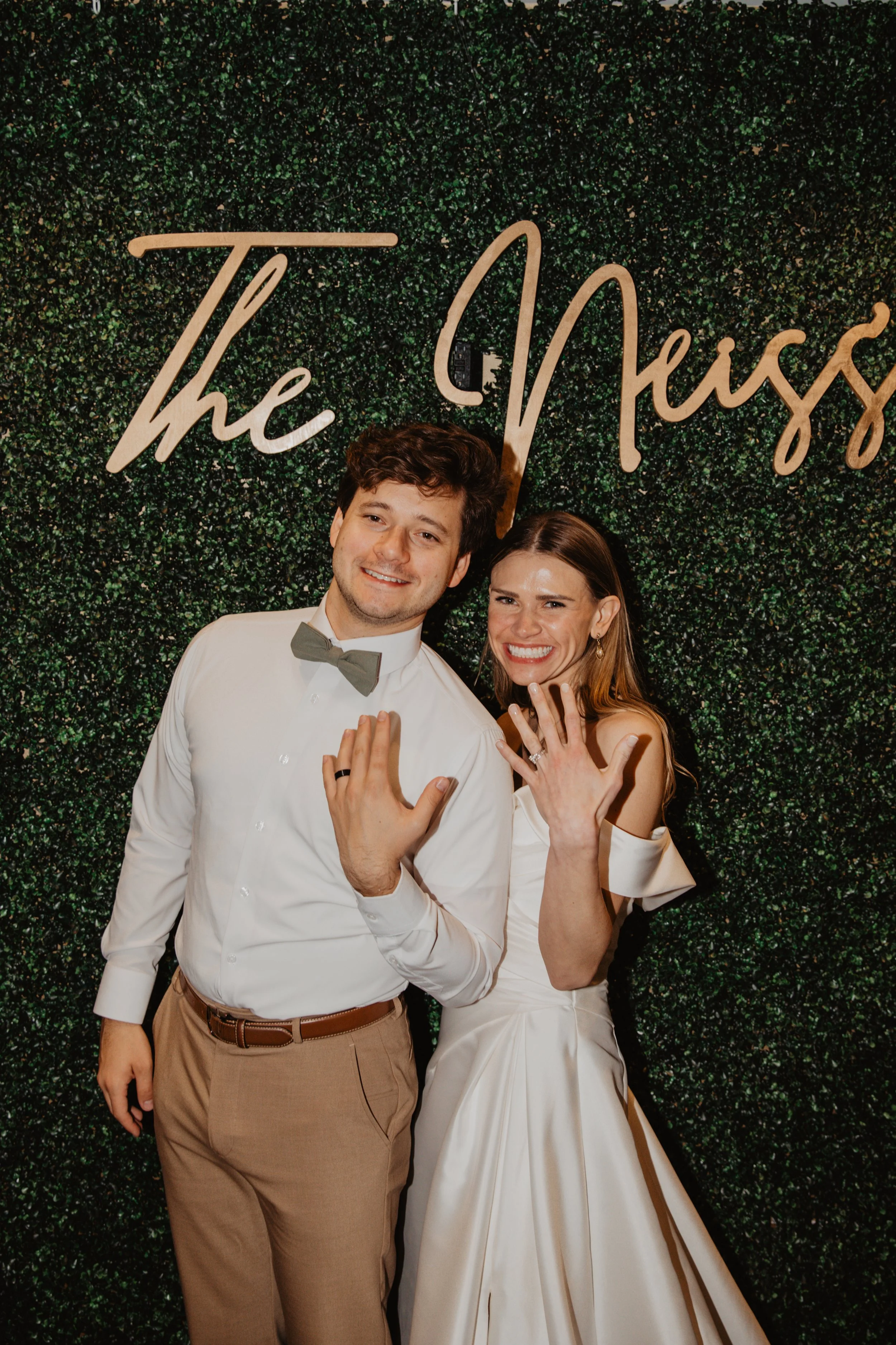 A happy couple showing off their wedding rings at a wedding celebration, standing in front of a green leafy backdrop with the partially visible words 'The' and 'Ness' in cursive gold lettering.