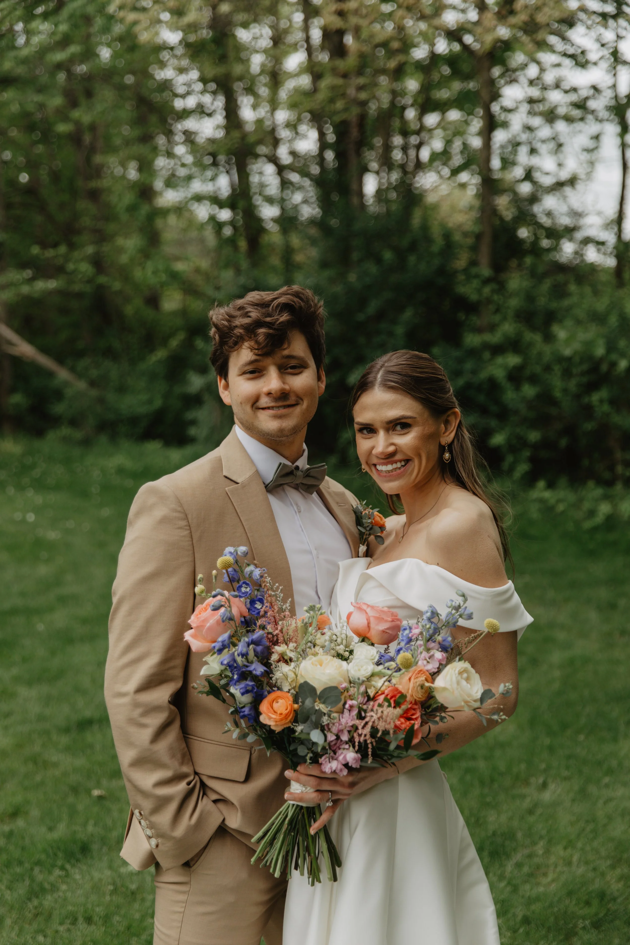 A newlywed couple smiling, standing outdoors on grass with trees in the background. The groom in a tan suit with a bow tie, the bride in a white off-shoulder wedding dress holding a large colorful bouquet.