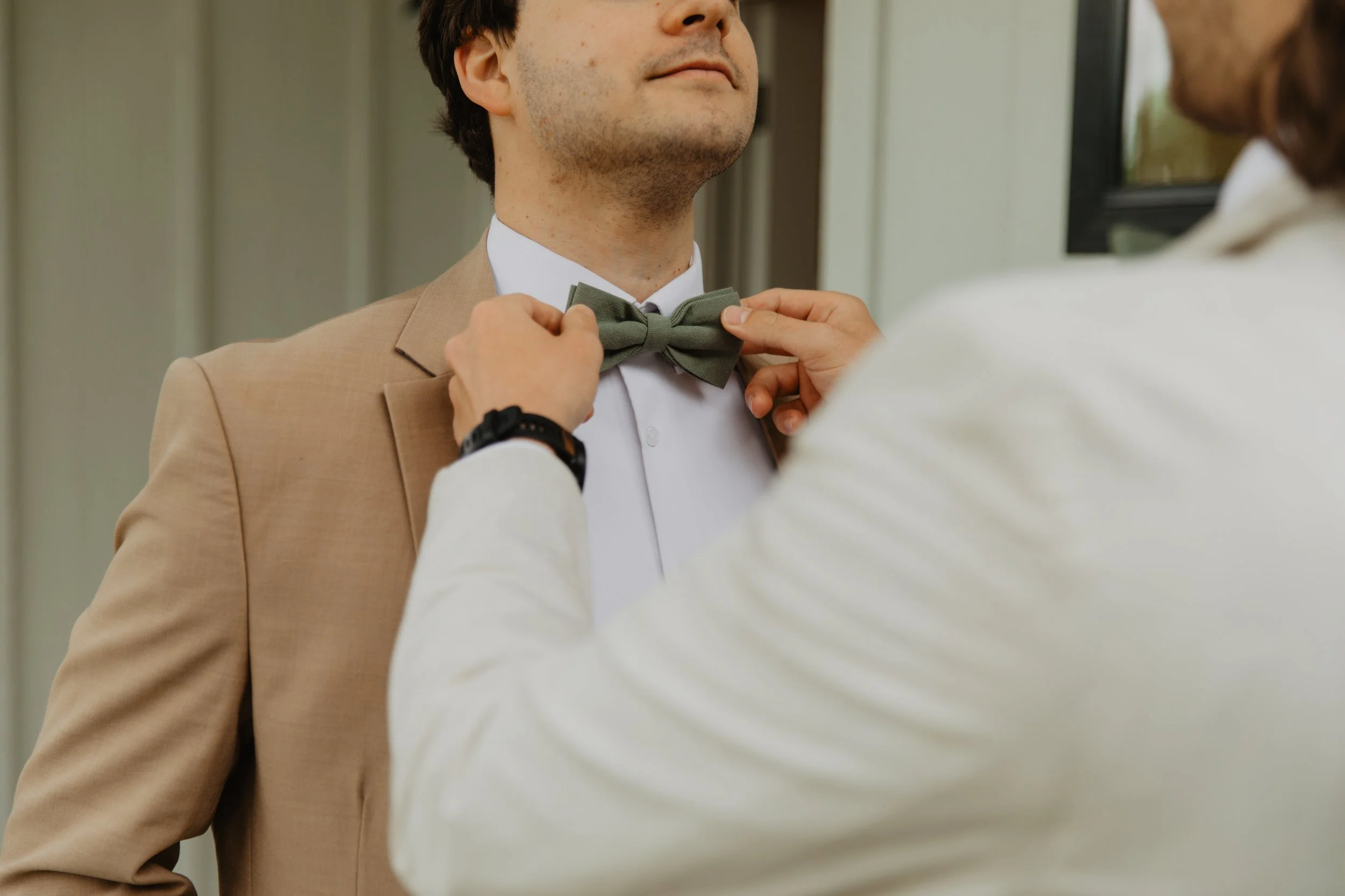 A man in a beige suit getting his green bow tie adjusted by another person in a white shirt.