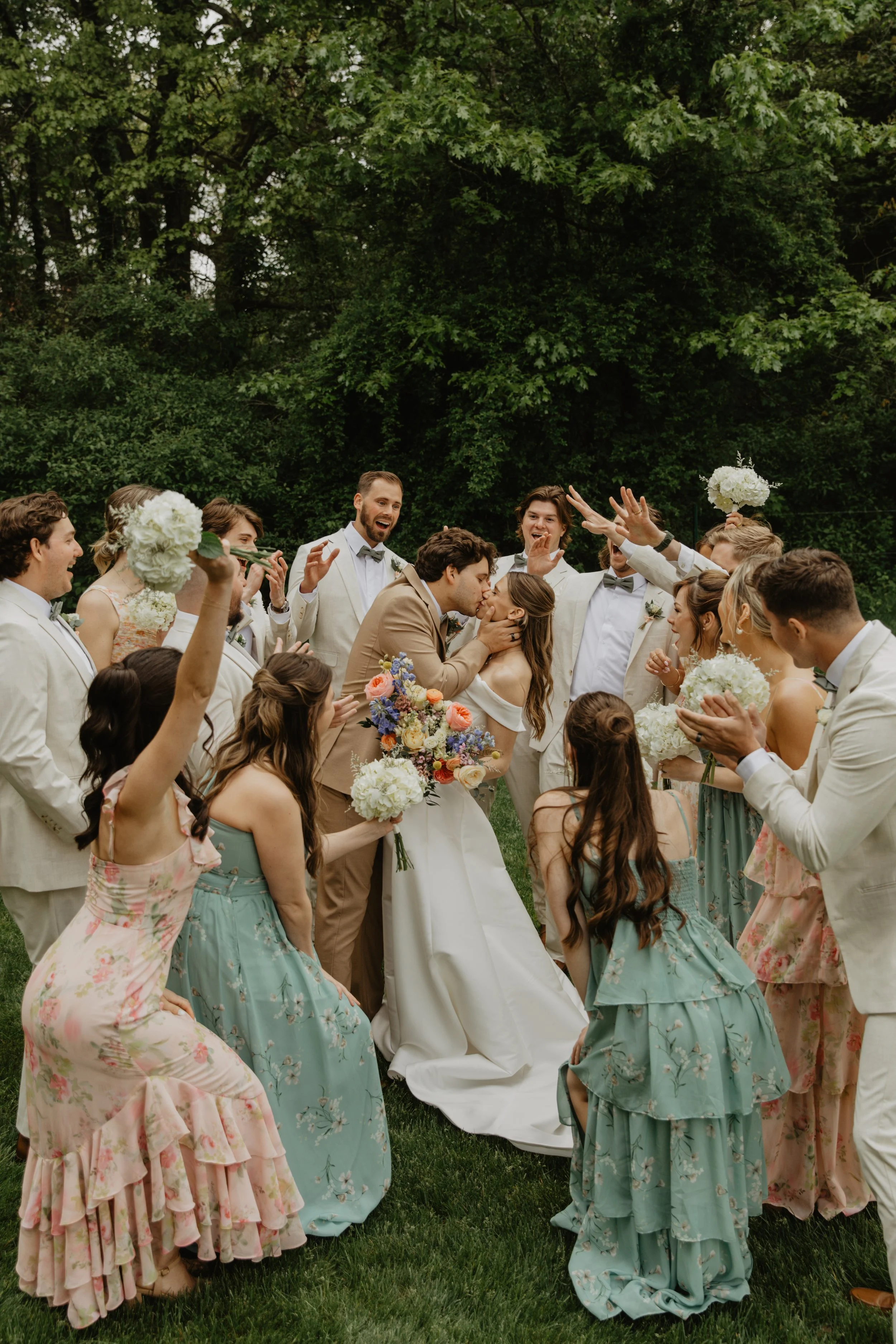 A wedding celebration outdoors with a bride and groom kissing in the center, surrounded by friends and family, some holding flowers and celebrating.