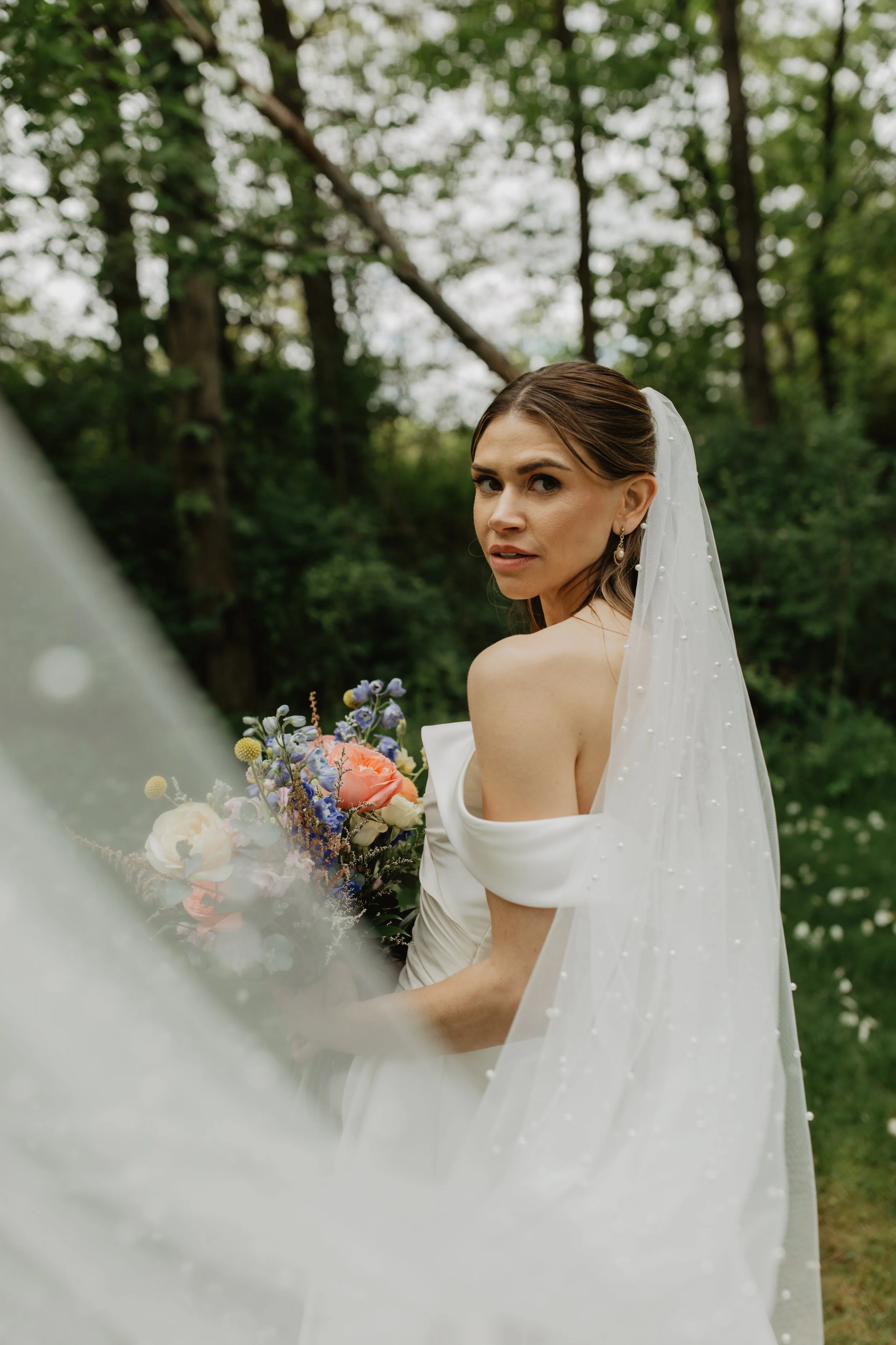 A bride standing outdoors in a forest, holding a colorful bouquet of flowers, wearing a white off-shoulder wedding dress and a veil, looking over her shoulder.